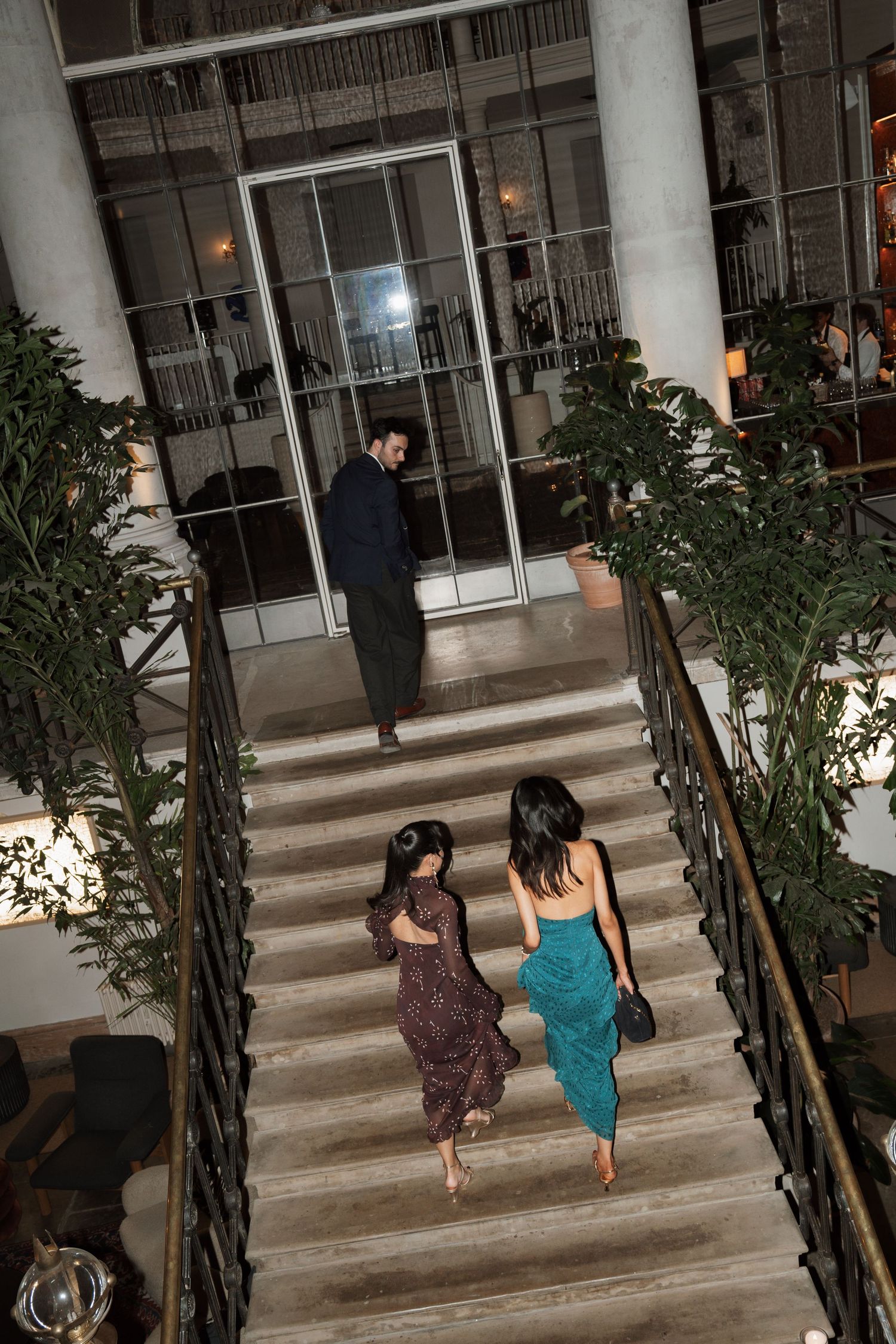 Two barefoot women in elegant dresses climb a wide stone staircase toward a glass-doored entrance where a man in dark clothing waits.