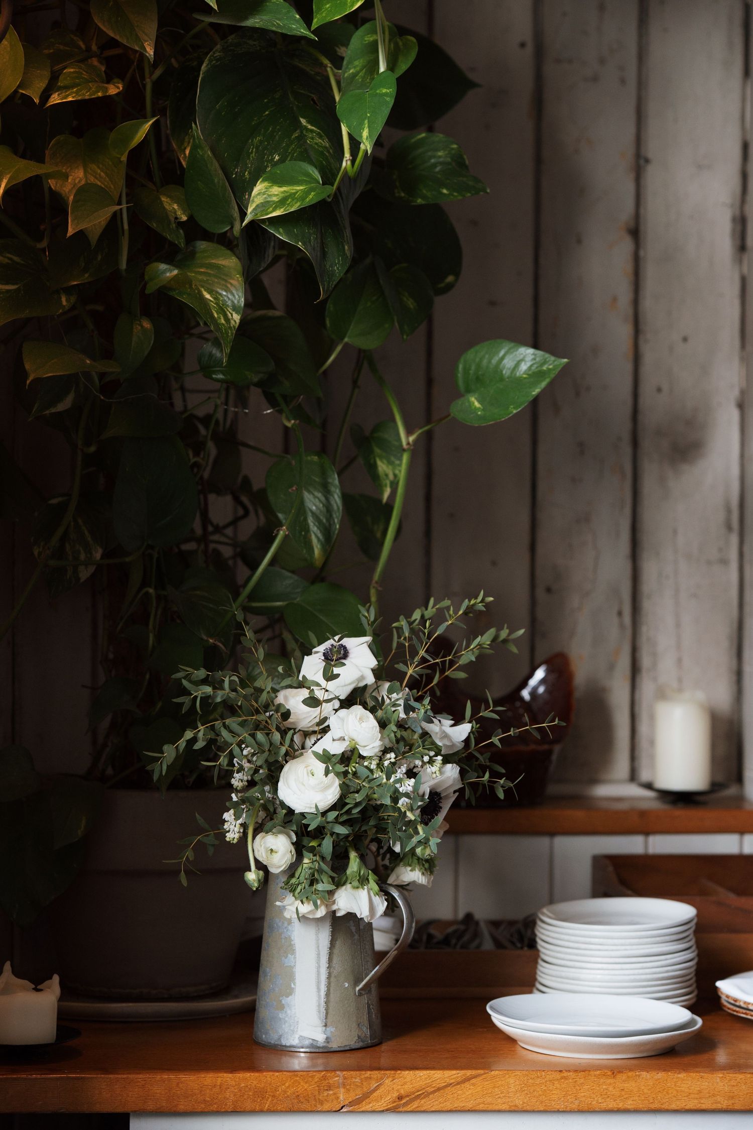 White floral arrangement in rustic vase sits on wooden shelf with stacked plates against grey wall.