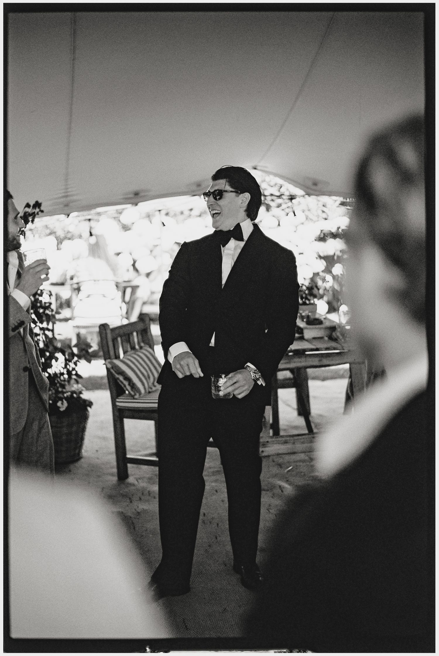 Man in tuxedo and bow tie wearing glasses stands confidently under a tent at a formal outdoor event.