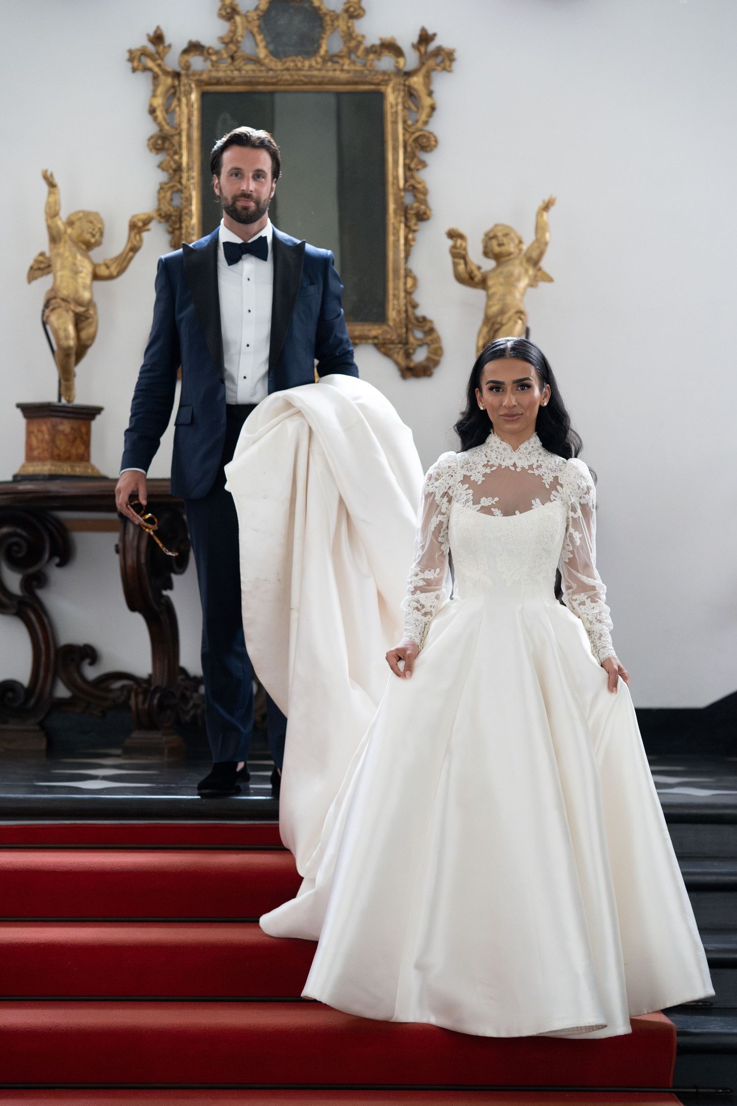 Wedding dress flowing dramatically on red carpet steps with ornate gold frames on wall.