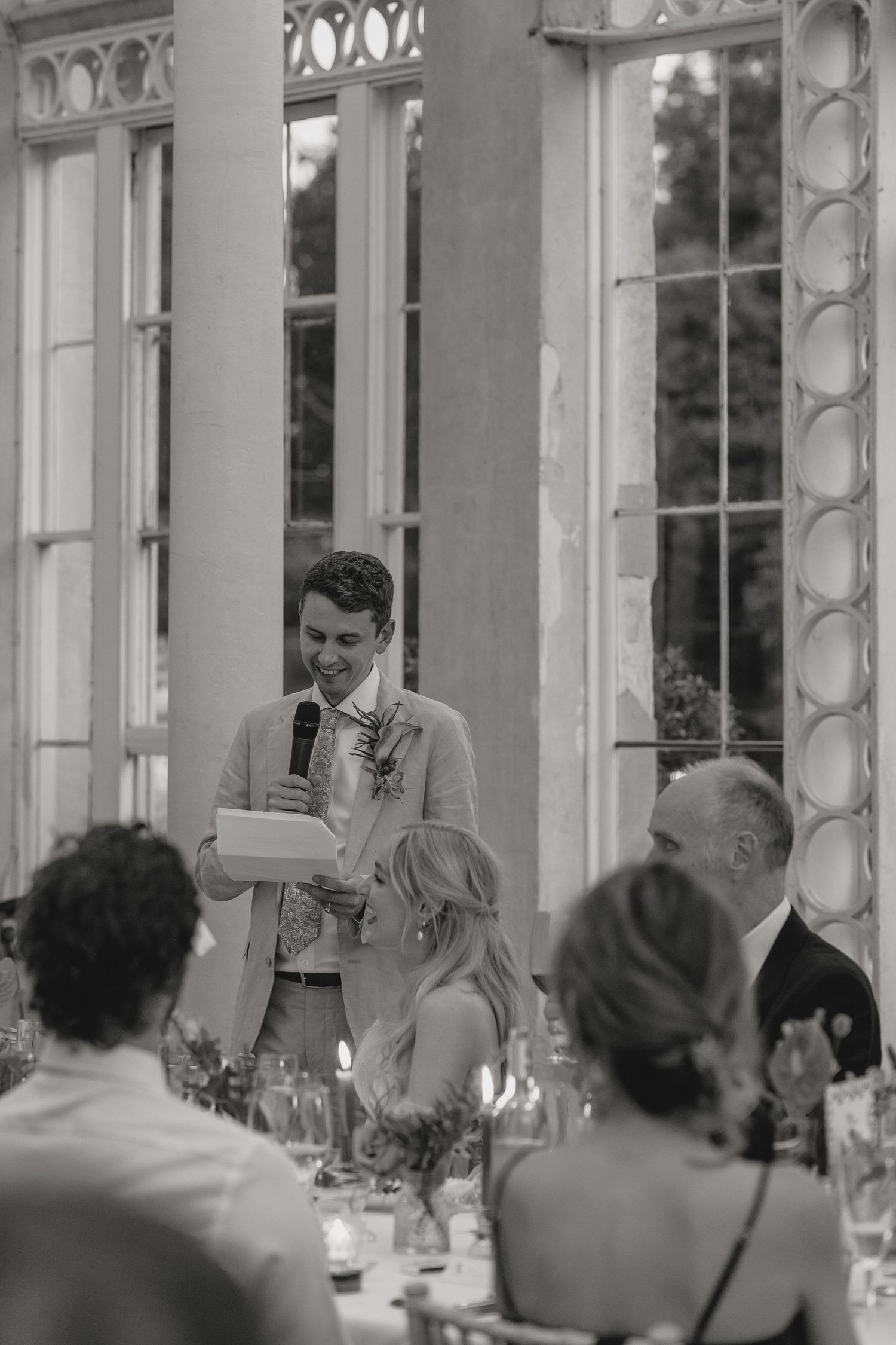 Black and white photo of wedding speeches in an elegant glass-walled venue.