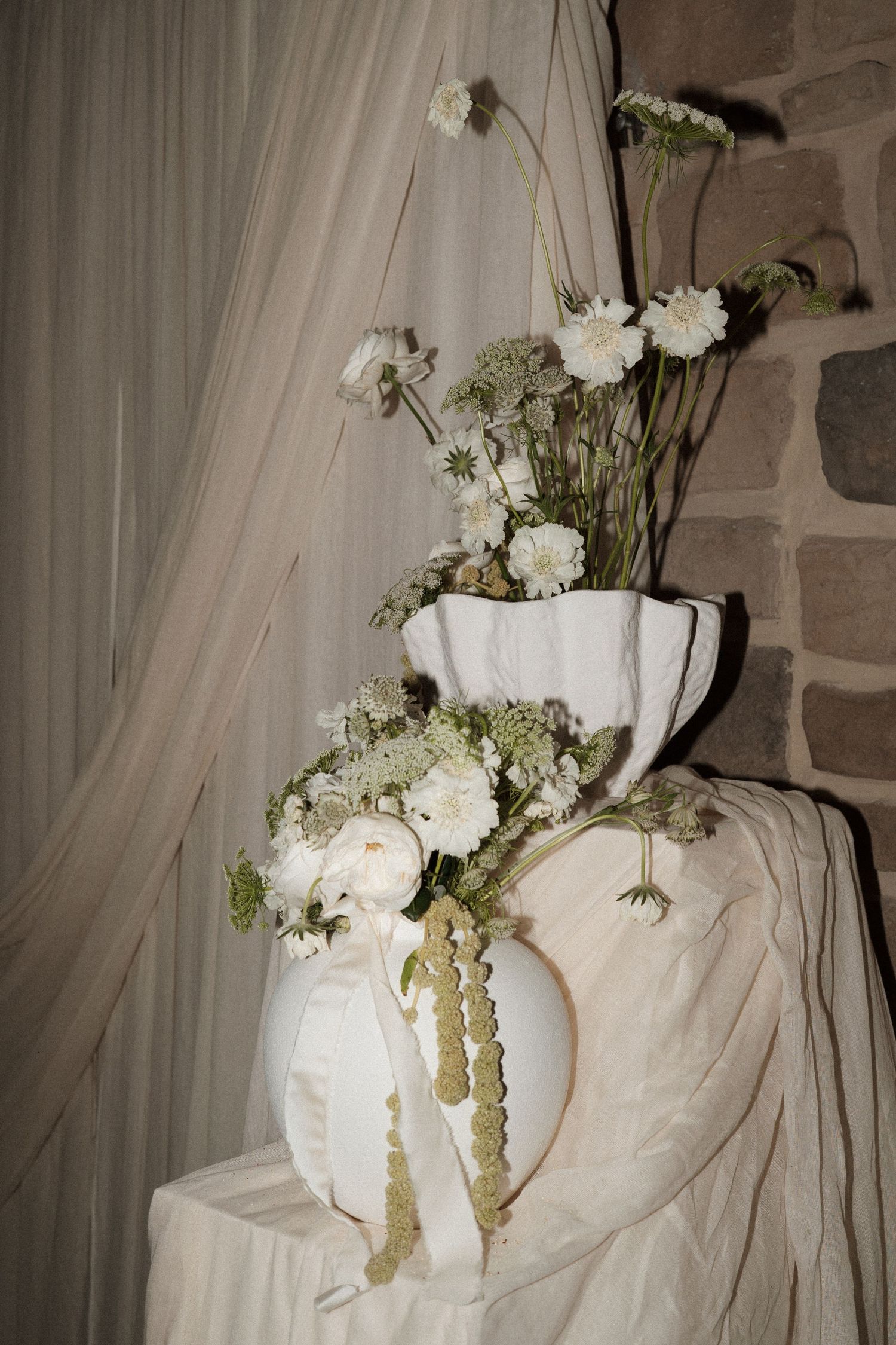 White roses and delicate flowers arranged in a textured ceramic vessel against stone wall.