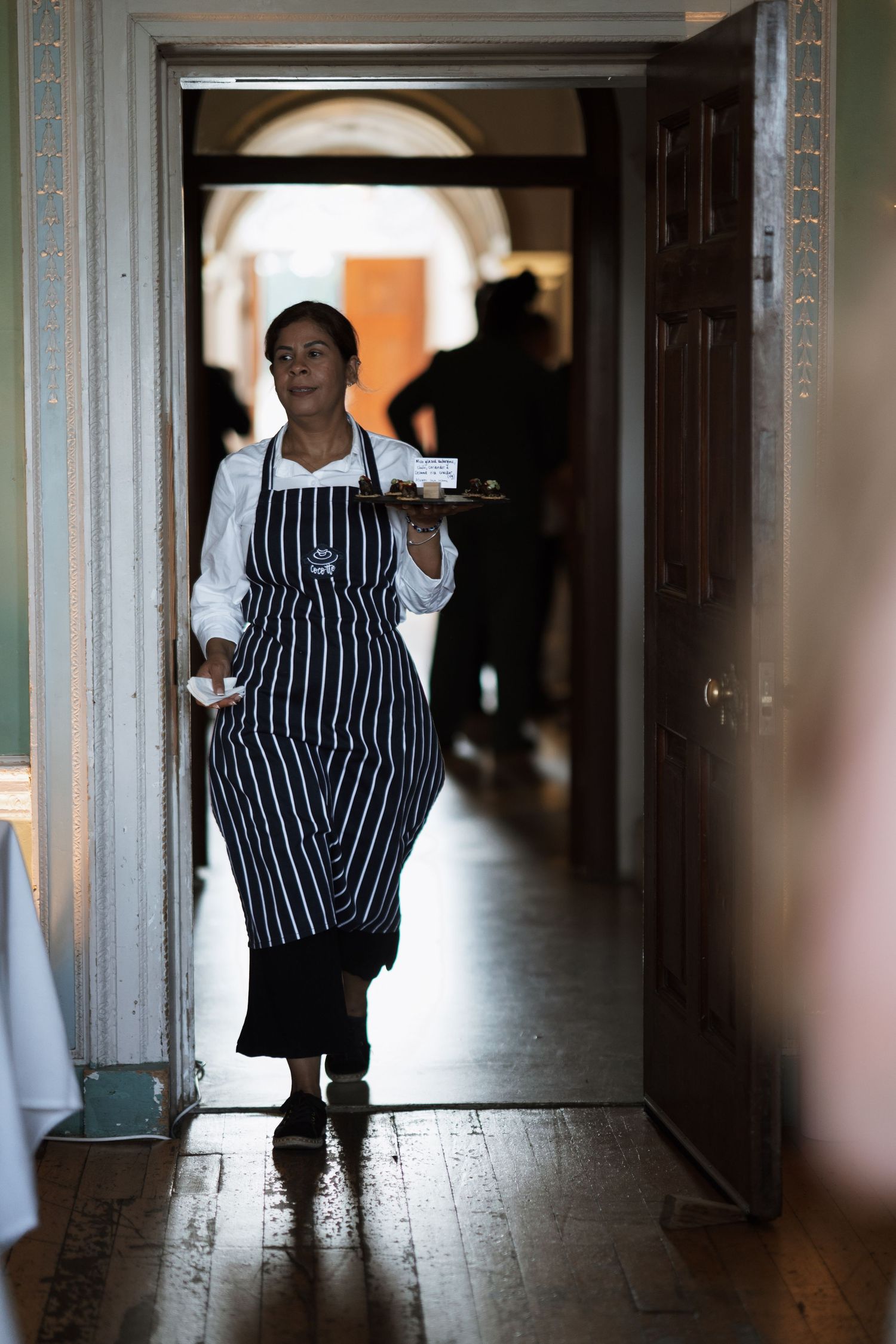 Server in striped apron carries tray through doorway at catered event.