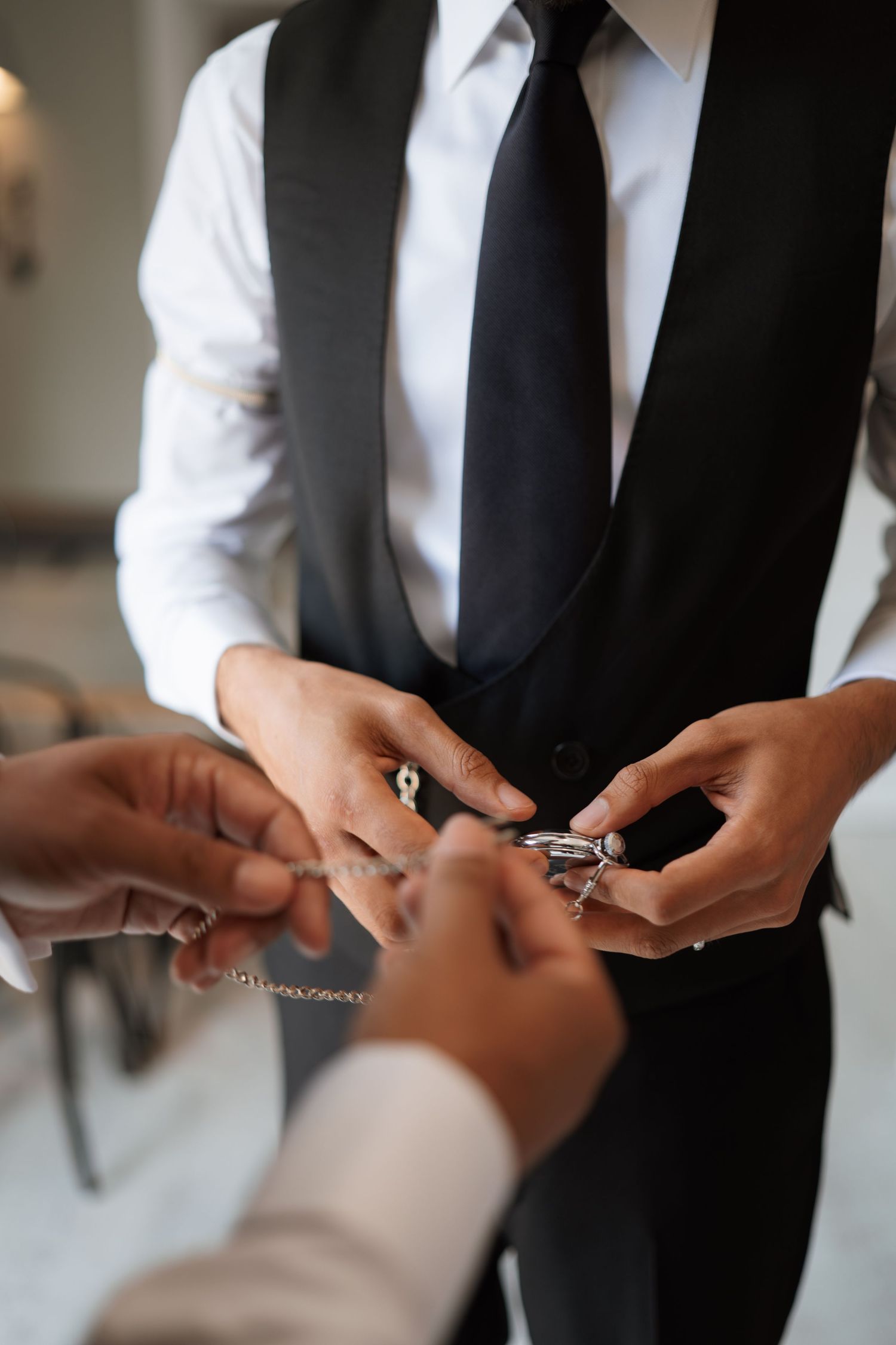 Hands exchanging jewelry or accessories, with person wearing formal black vest and tie.