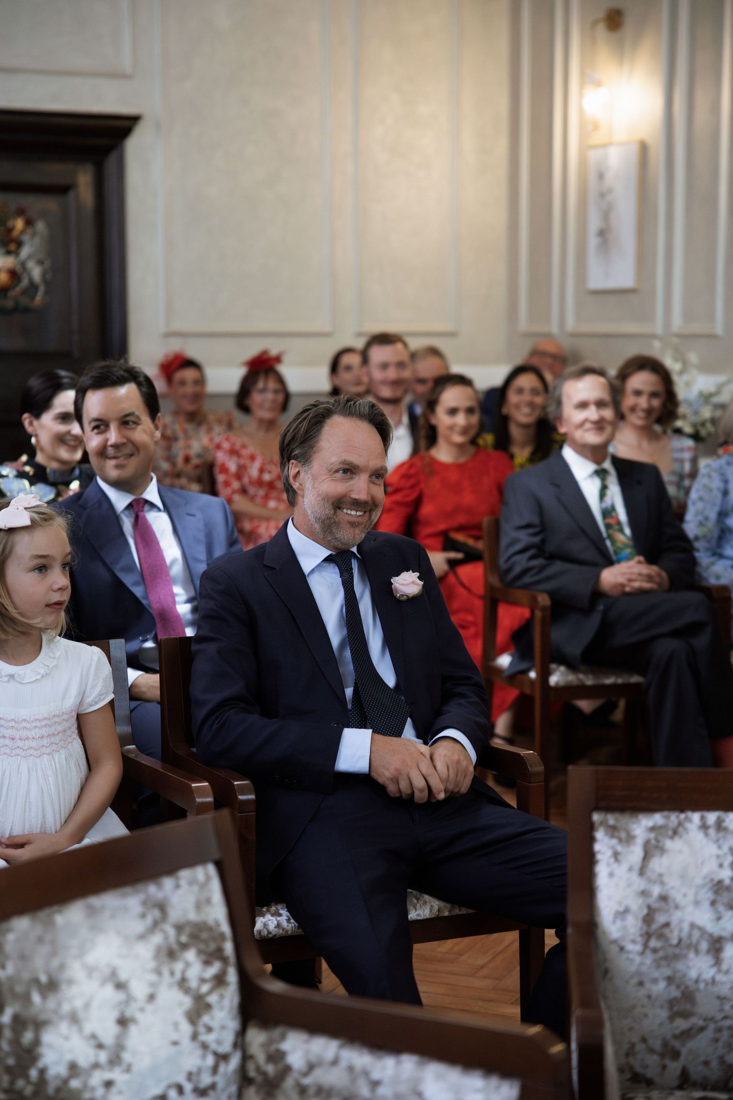 Wedding guests seated in rows at a ceremony venue with ornate white walls.