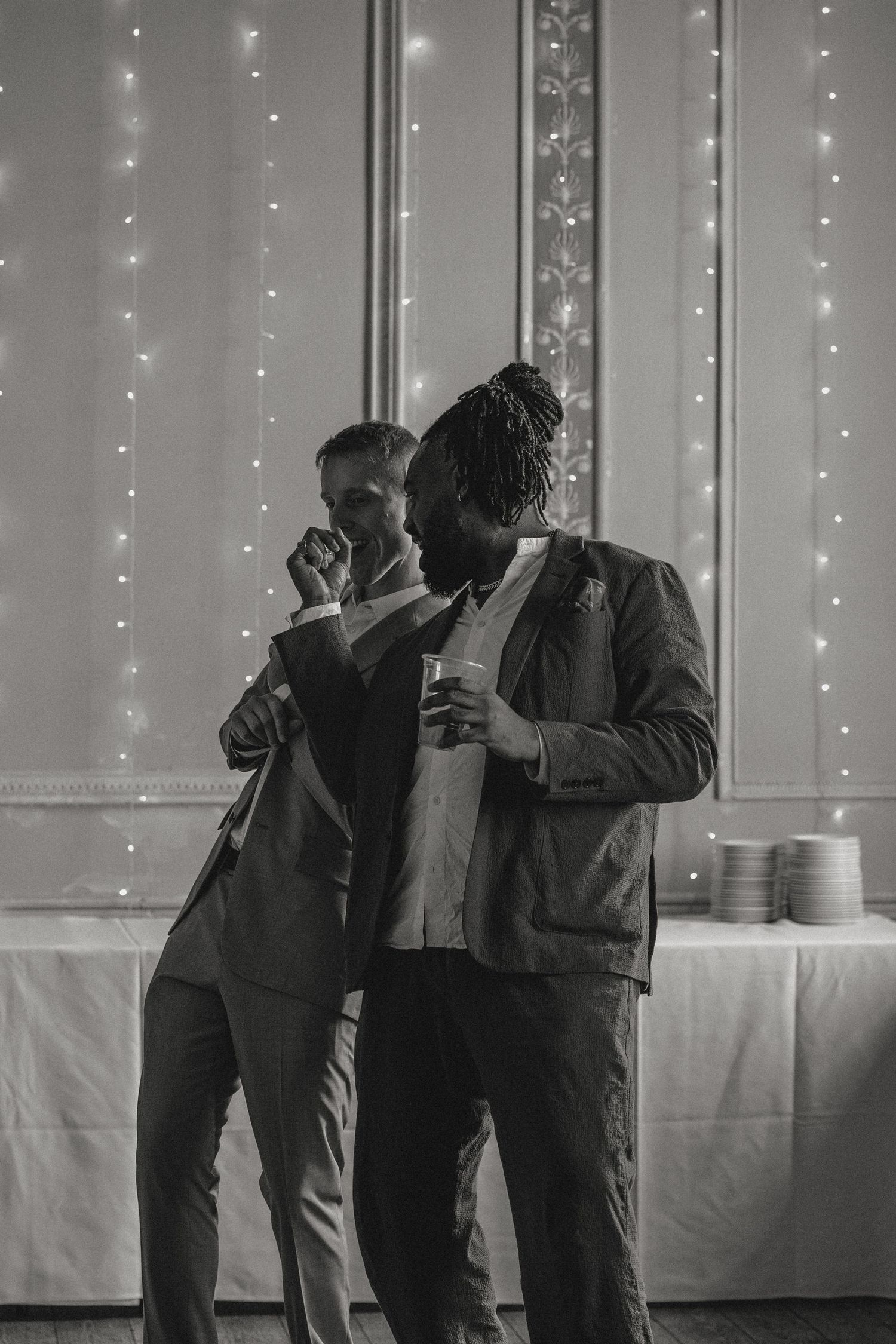 Two guests share drinks under twinkling fairy lights at evening event.