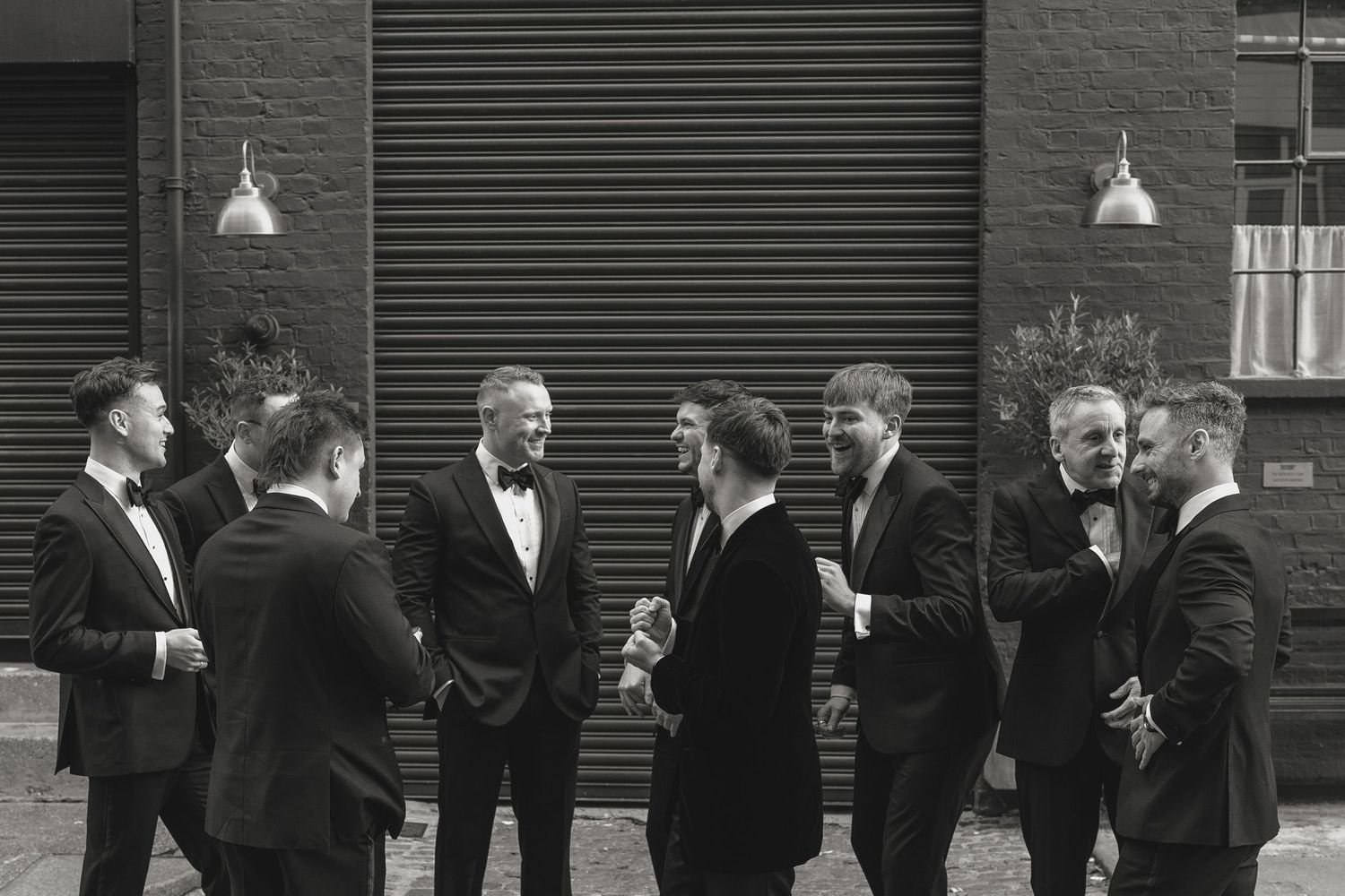 A group of people in formal attire gathered against an industrial wall with pendant lights.