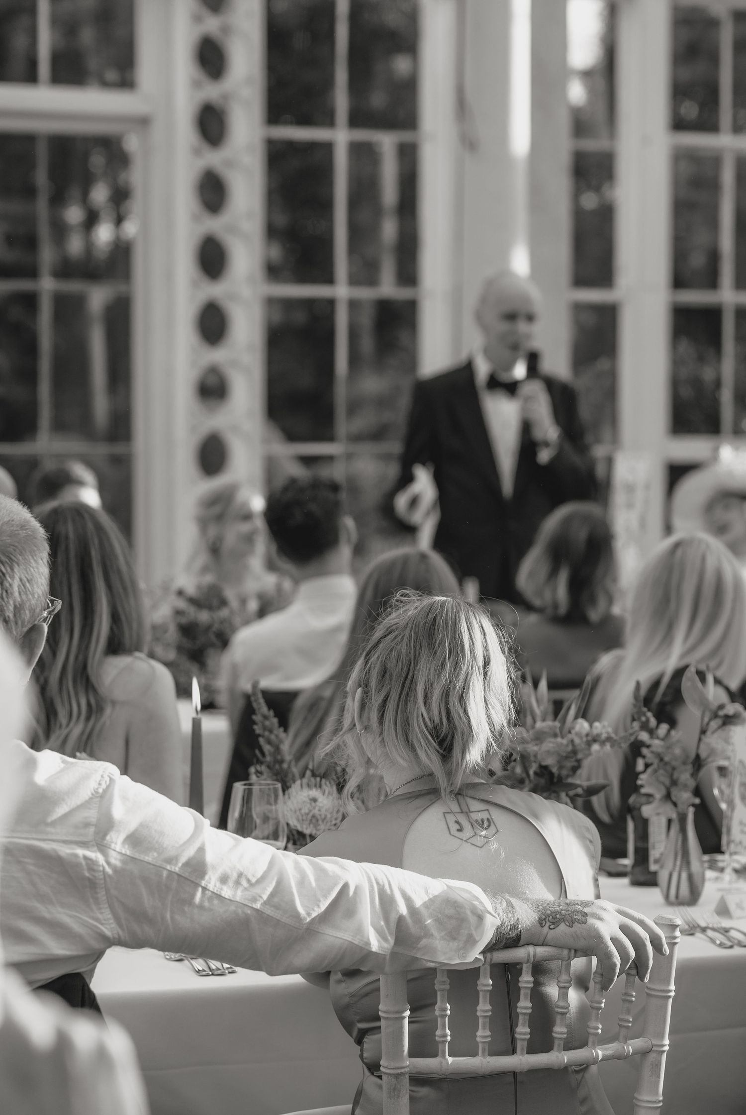 Wedding guests seated at long tables in a bright conservatory venue.