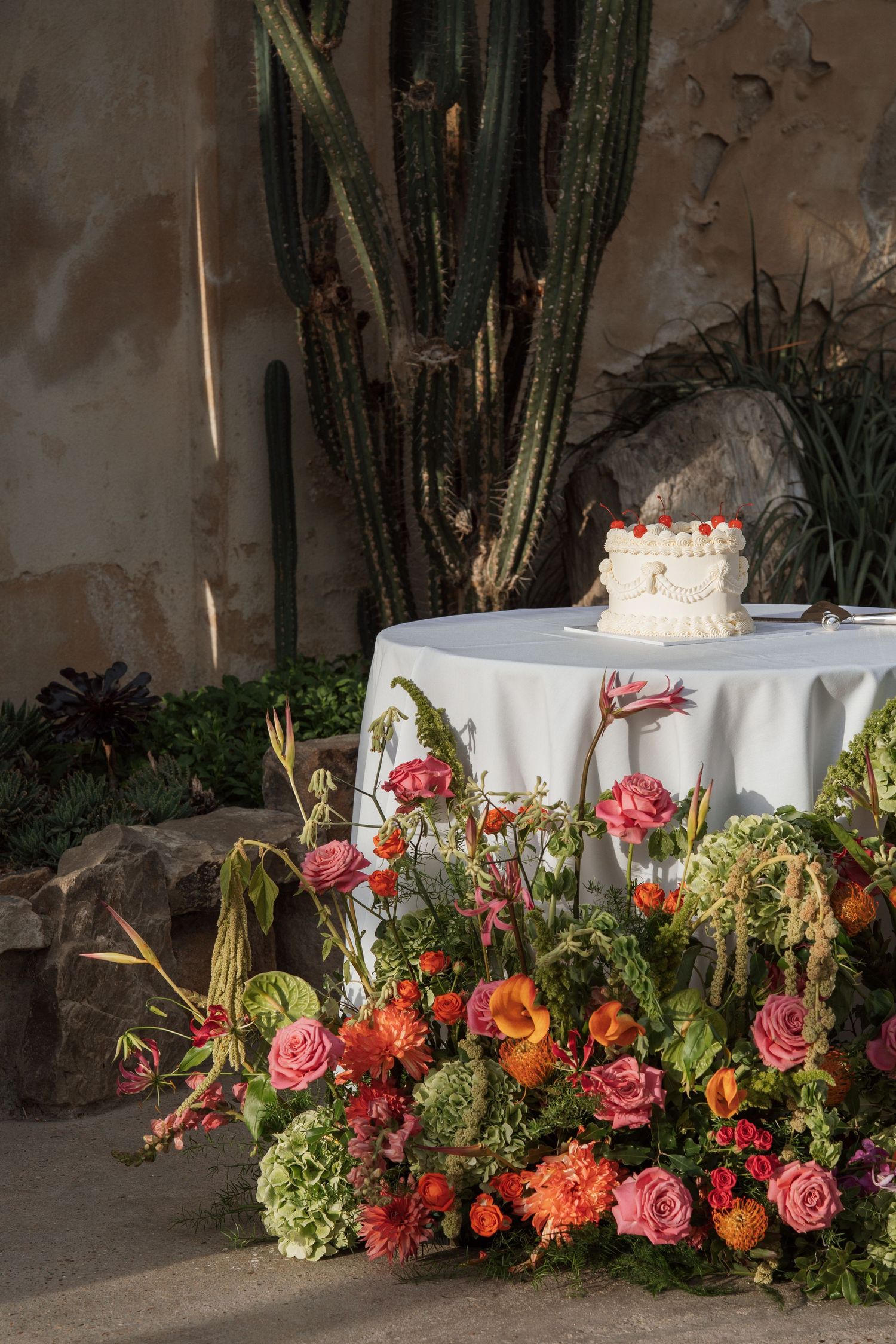 Wedding cake on draped table with vibrant pink and orange floral arrangements, set against rustic wall with tall cacti.