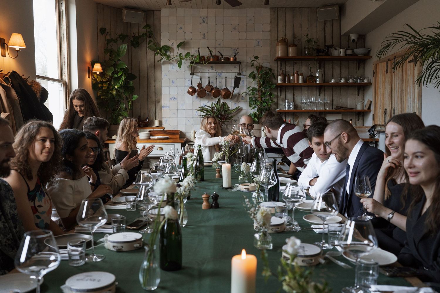 Guests gathered around a long dining table set with candles and greenery in a rustic restaurant for a festive dinner party.