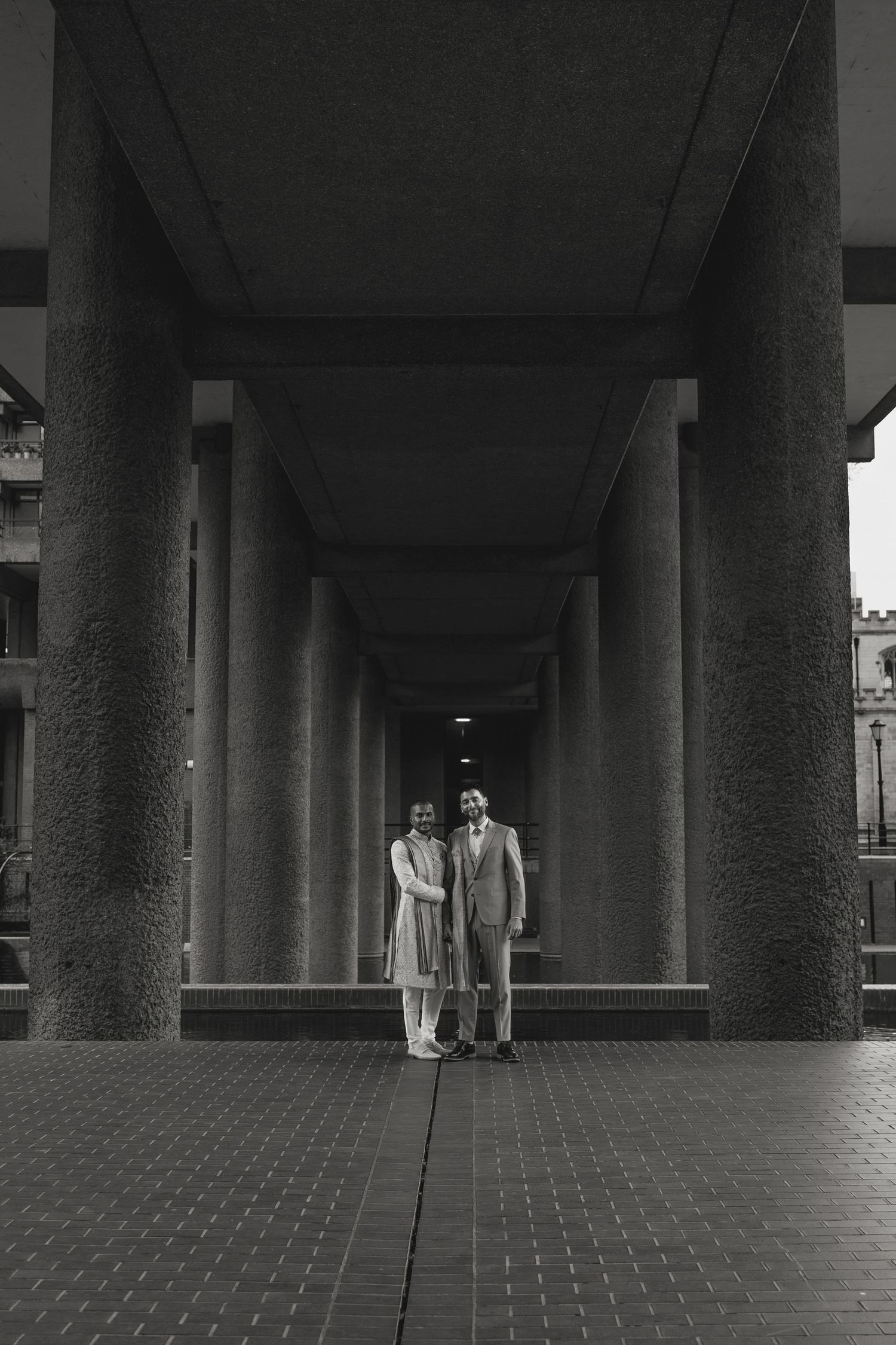 Black and white photo of a couple standing between large columns in an elegant architectural corridor.