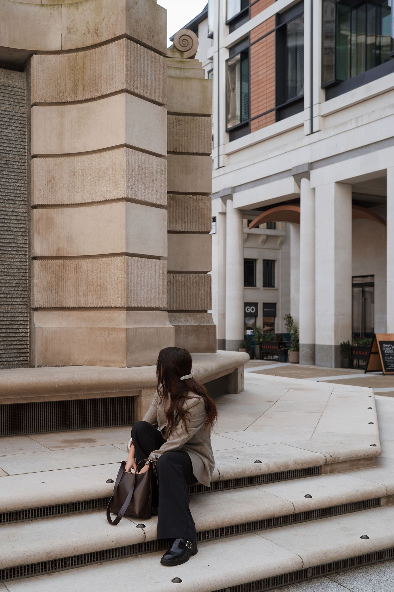 Woman in beige coat sitting on stone steps outside a classical building, holding a brown leather bag.