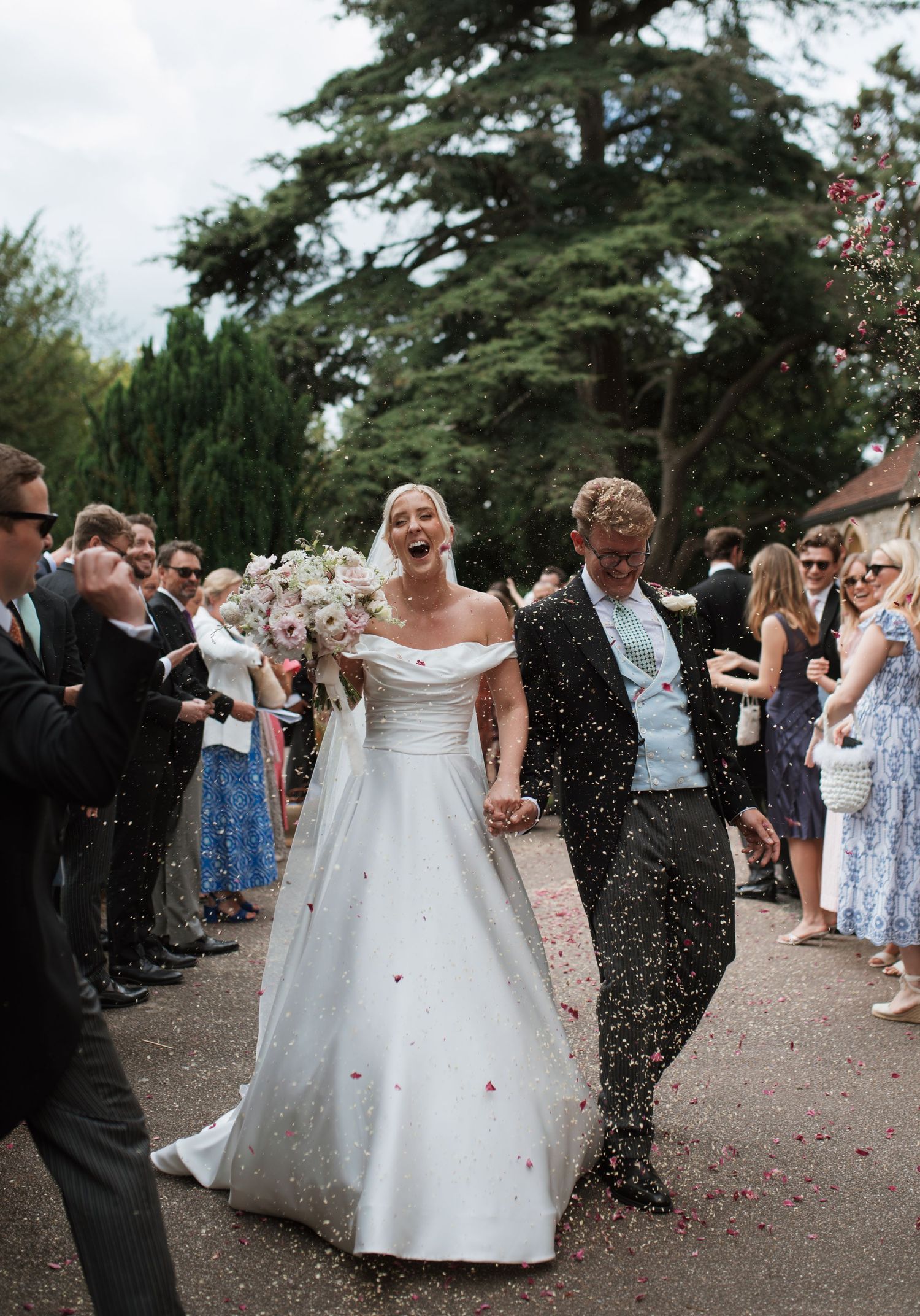 Joyful bride in white off-shoulder gown and groom walk through confetti shower, guests cheering along outdoor wedding path.