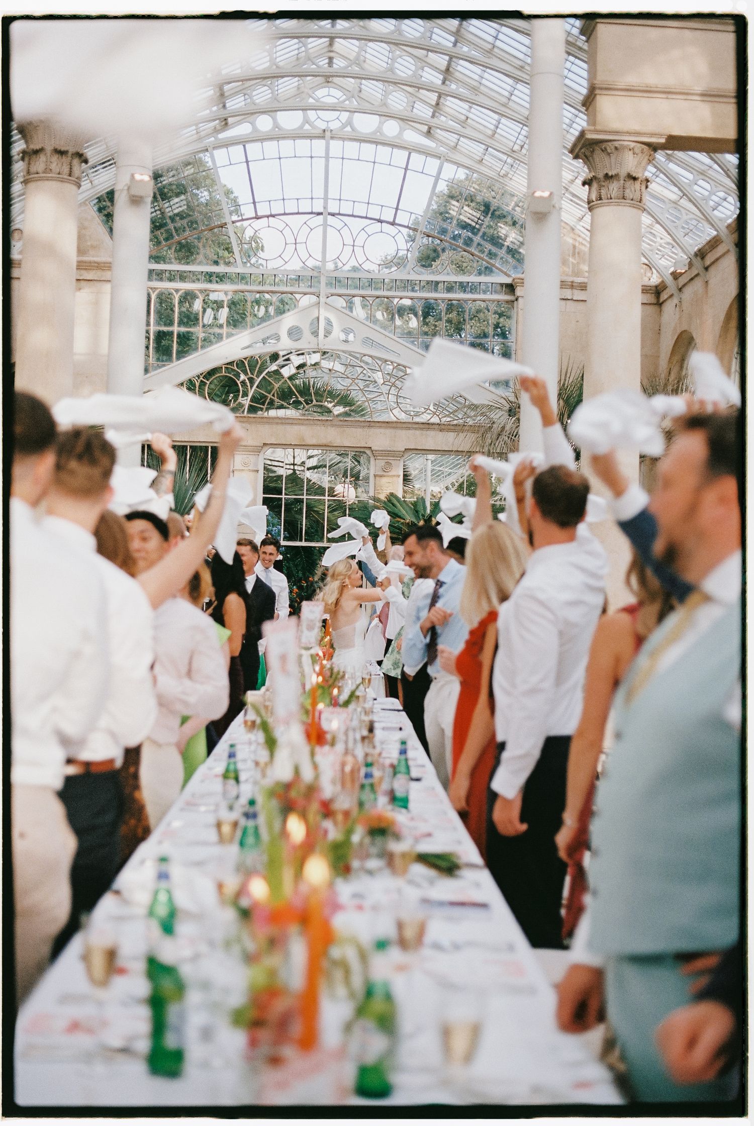 Elegant wedding reception in a glass conservatory with guests waving white napkins along a long decorated table.