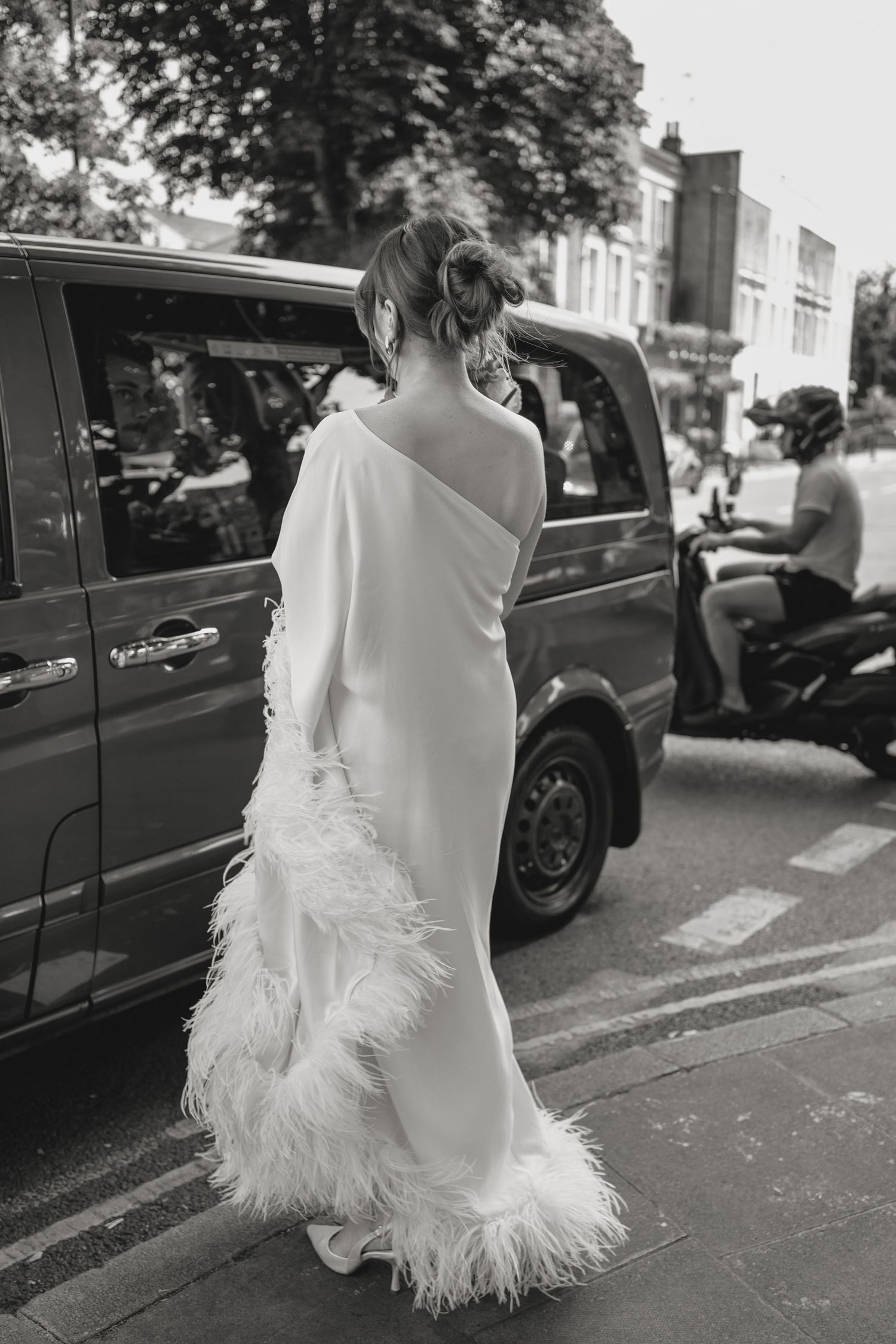 A bride in an elegant white dress with feather details standing next to a car on a city street.