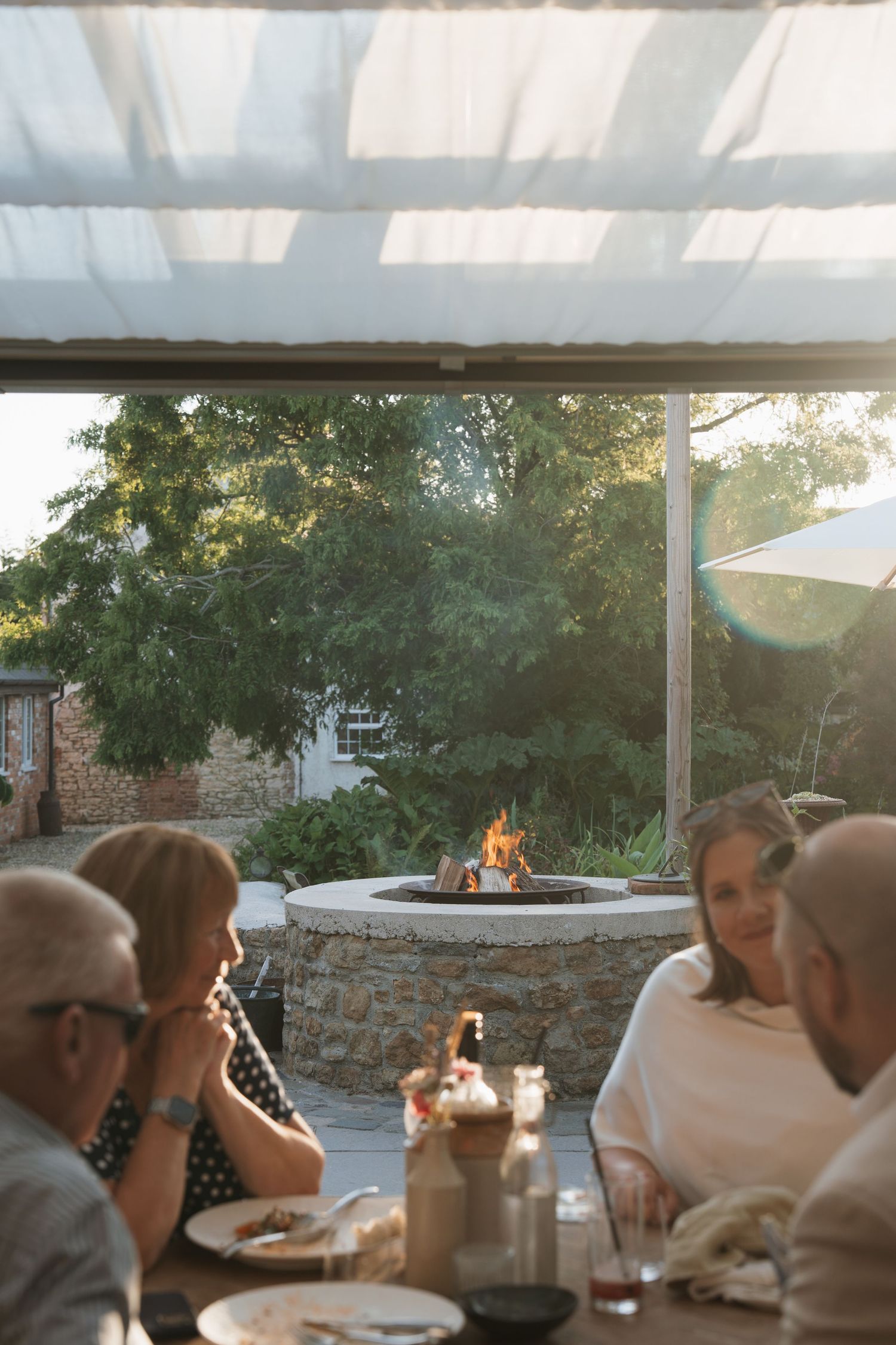 Group dining outdoors under pergola with stone fire pit glowing in background at sunset.