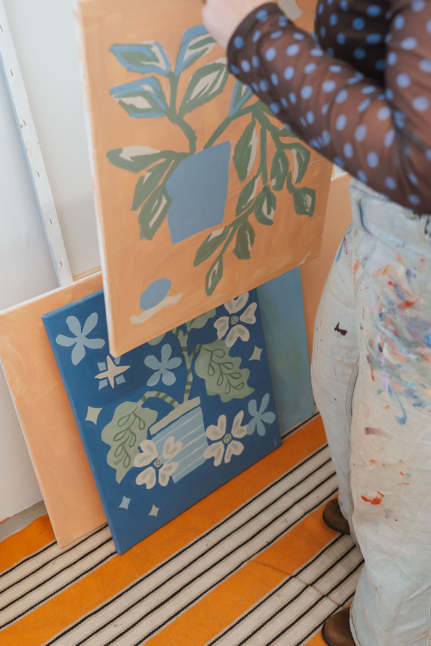 Artist in paint-splattered pants arranging botanical paintings in blue and orange tones against a studio wall.