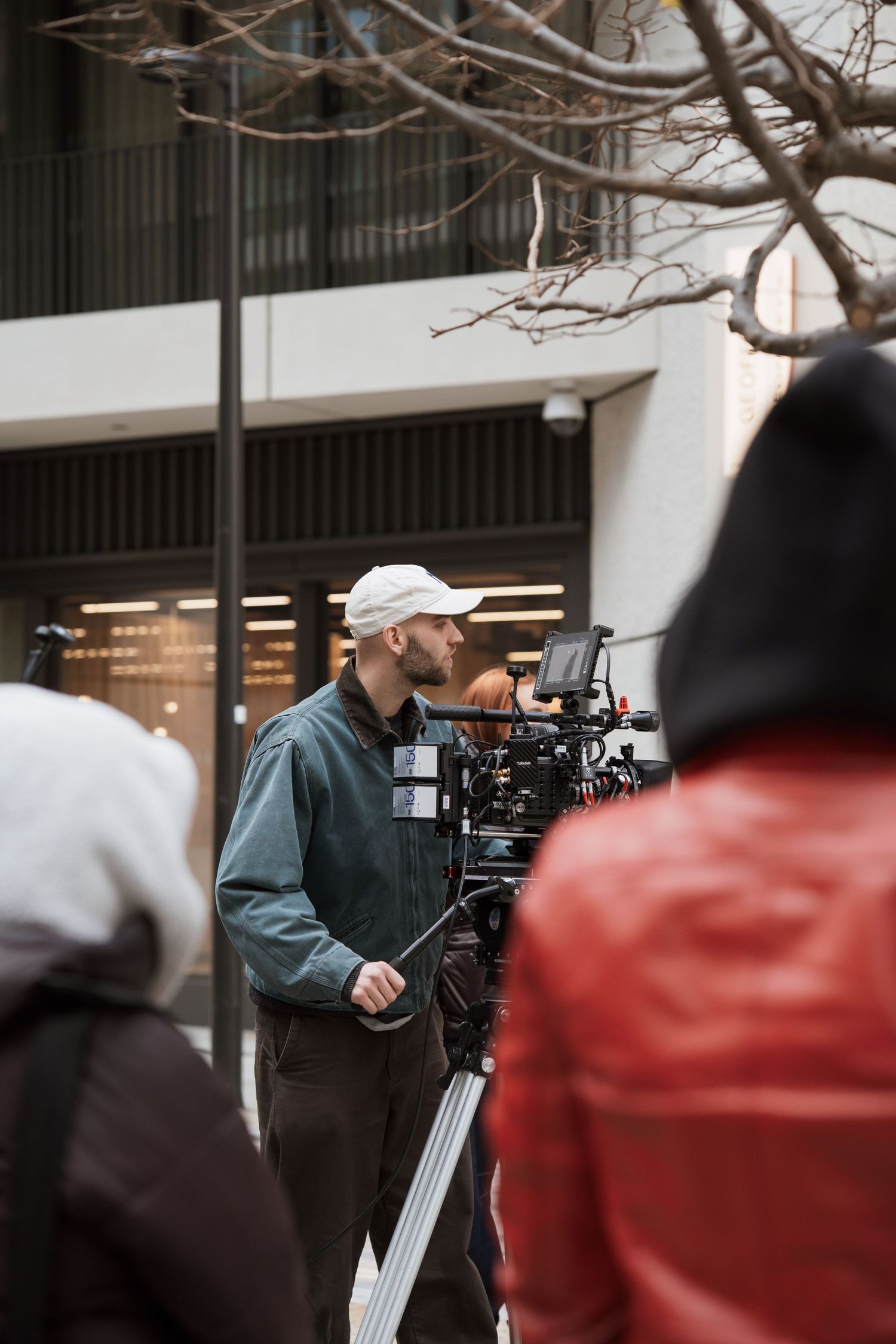 Cameraman in white cap operating professional film camera outdoors on urban street during video production shoot.
