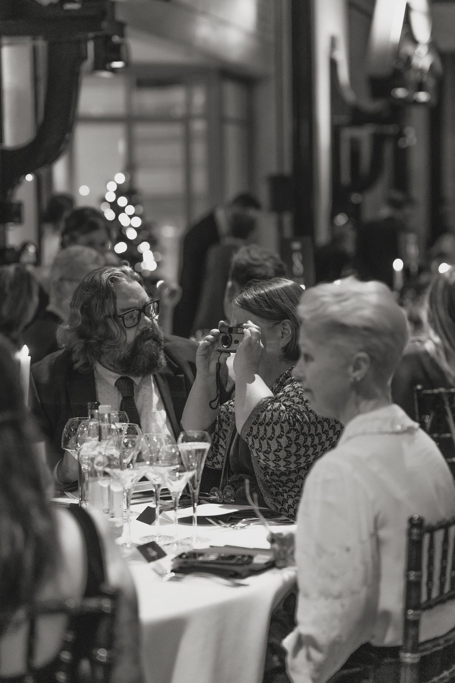 Elegant dinner party scene with guests seated at tables in a dimly lit restaurant setting.