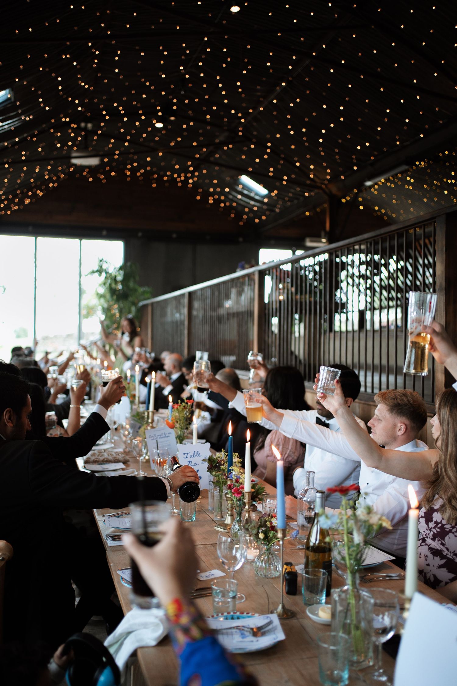 Wedding guests raise their glasses in celebration during a reception dinner under twinkling lights.