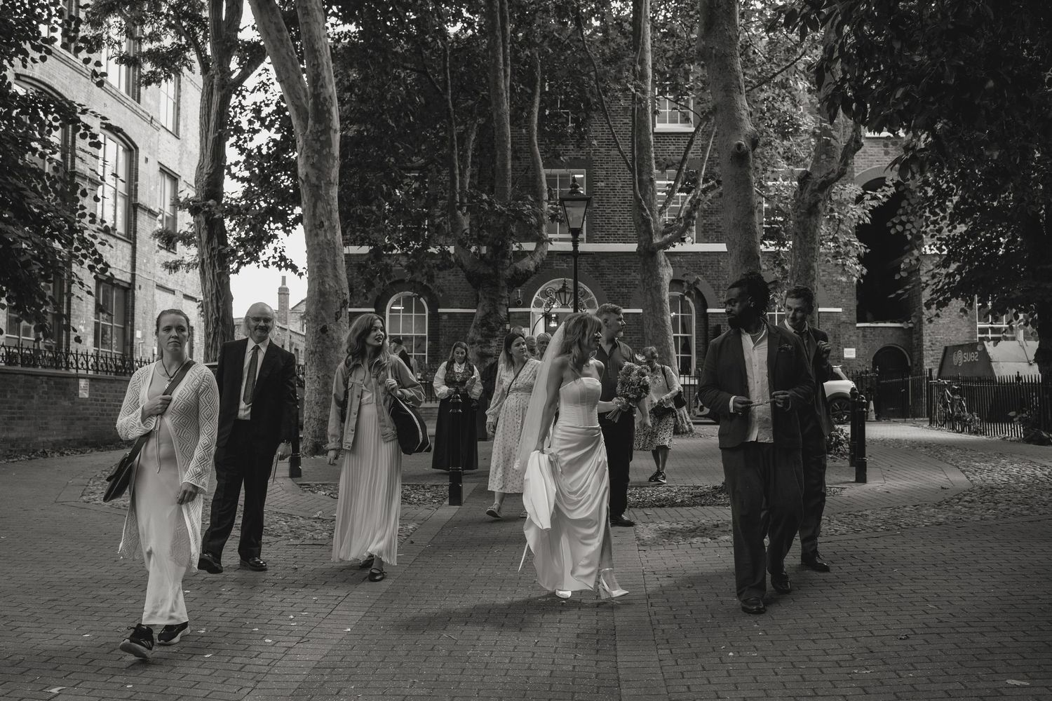 Wedding party walking through a tree-lined path in formal attire.