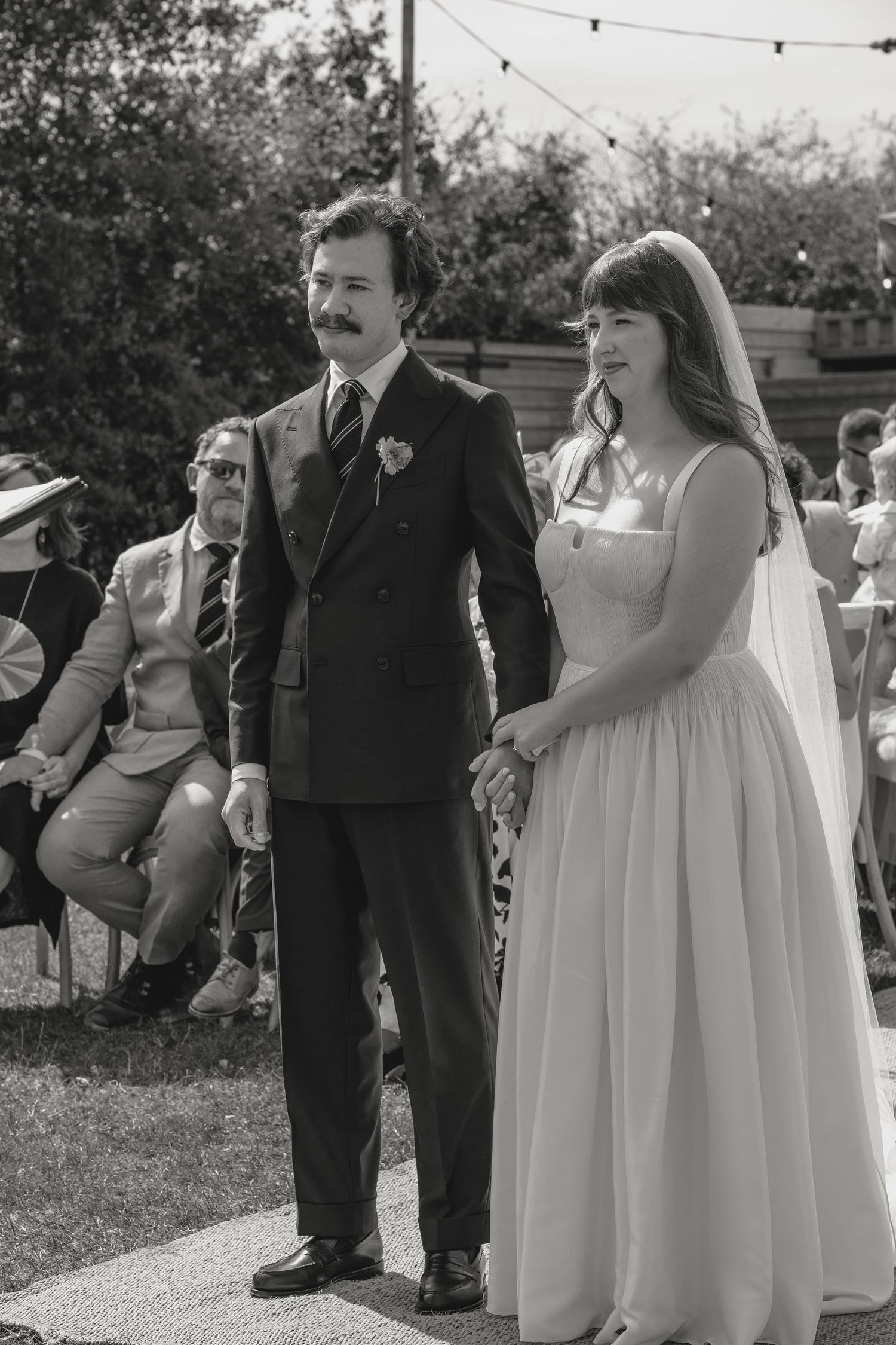 Wedding party members in formal attire stand together under string lights outdoors.