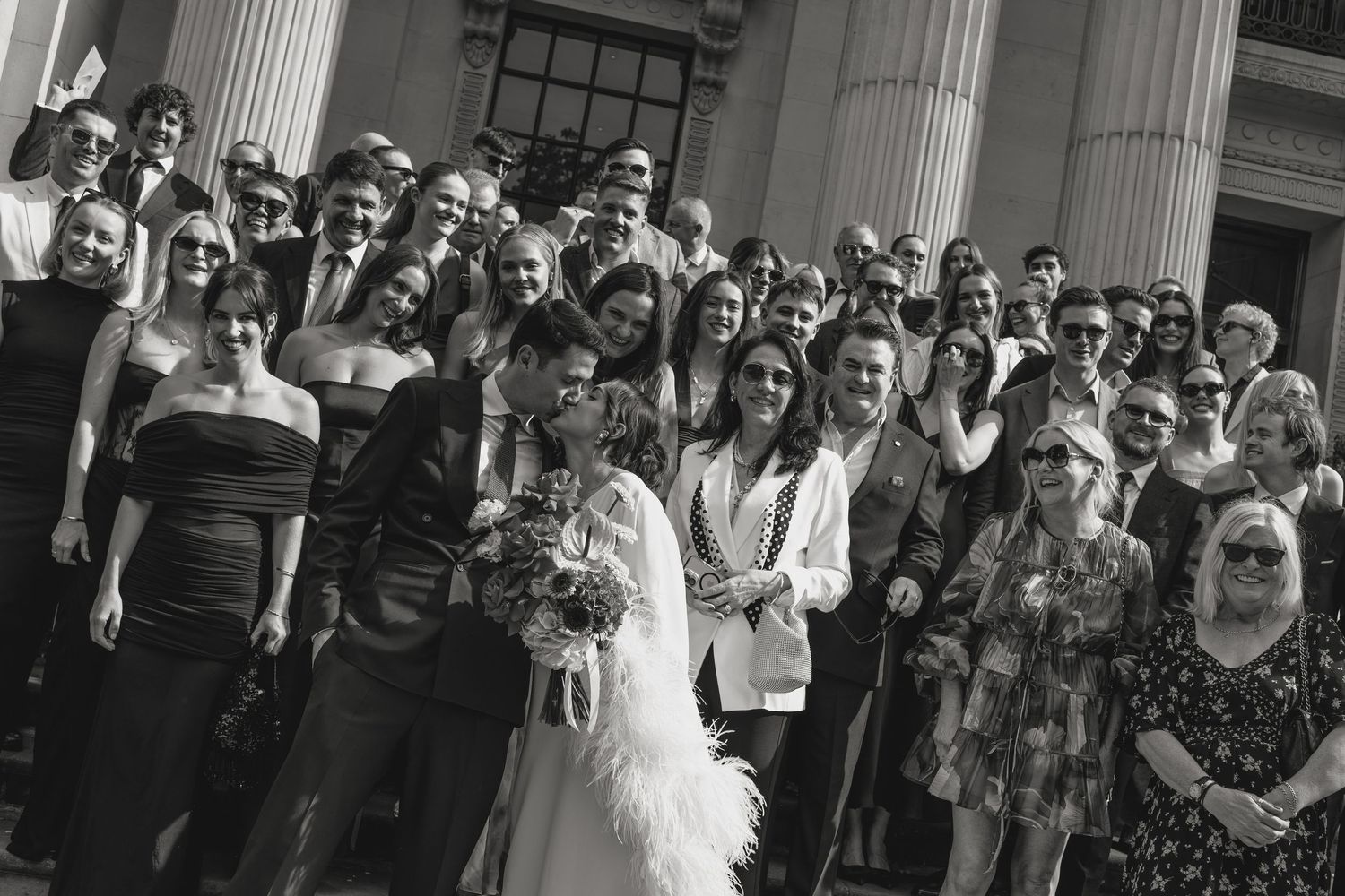 Large wedding party gathered on steps of historic building with white columns.