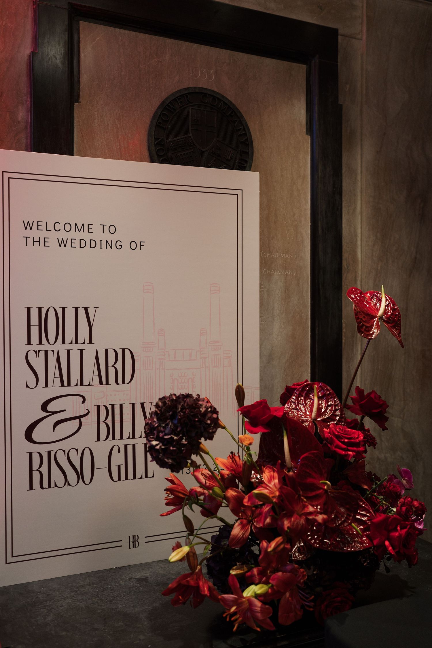 Wedding welcome sign with red floral decorations displayed against a dark wall.