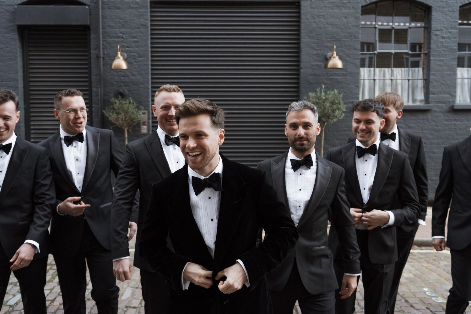 Groomsmen in black tuxedos pose together against industrial brick backdrop.