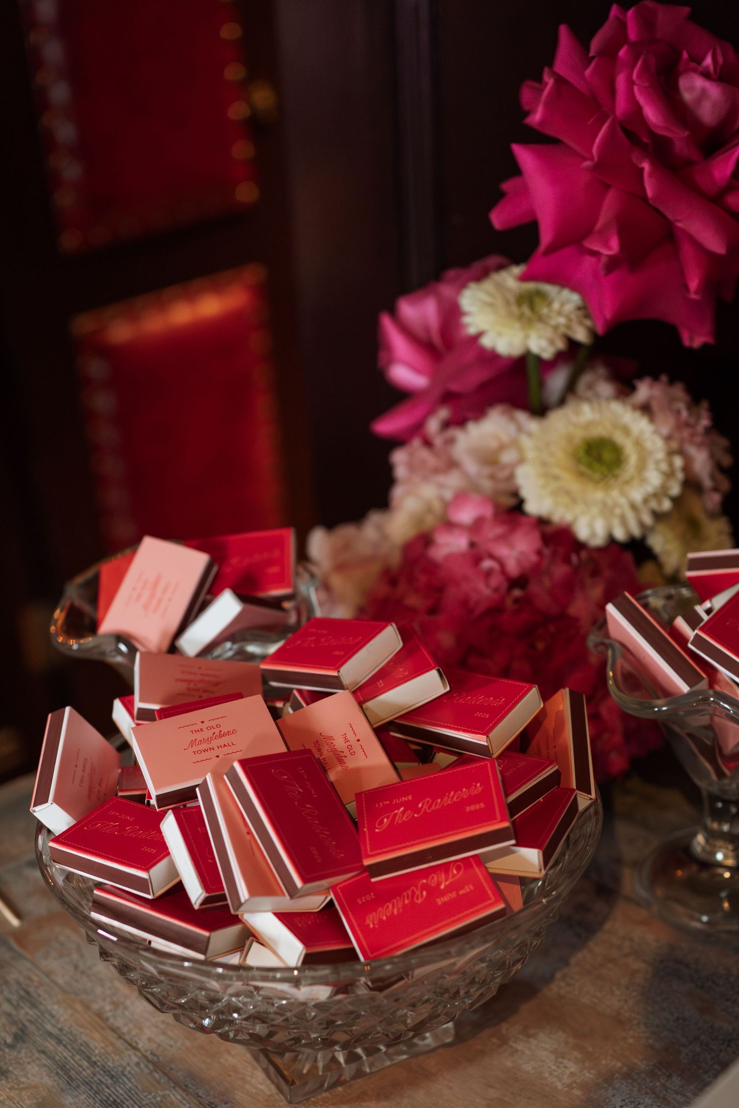 Crystal bowl filled with red matchbooks and pink flowers on wooden surface.
