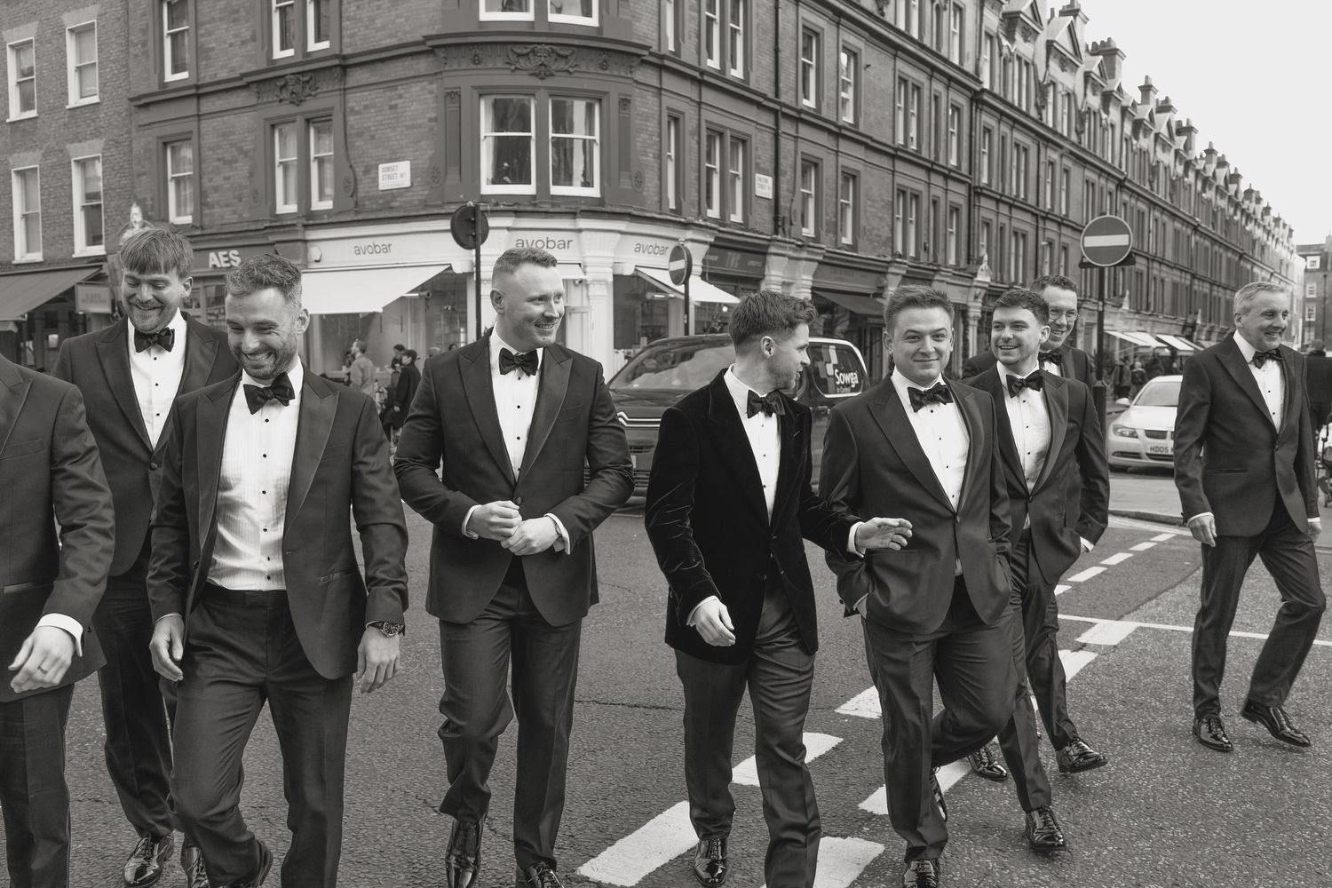 A group of formally dressed men in tuxedos walking down a city street in black and white.