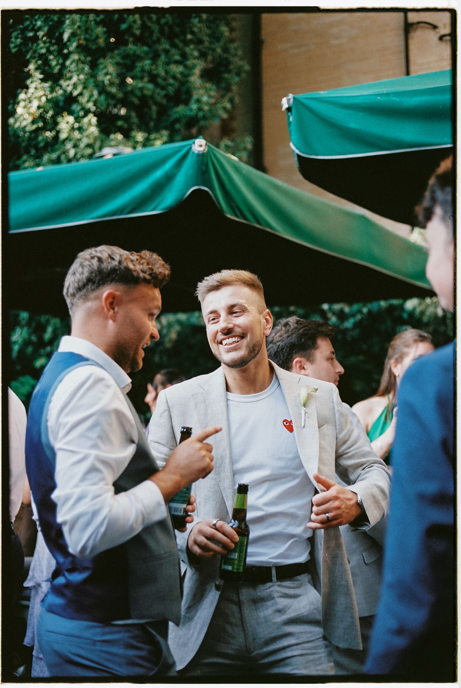 Two men in smart casual attire laugh and chat outdoors at a social event, holding green beer bottles.
