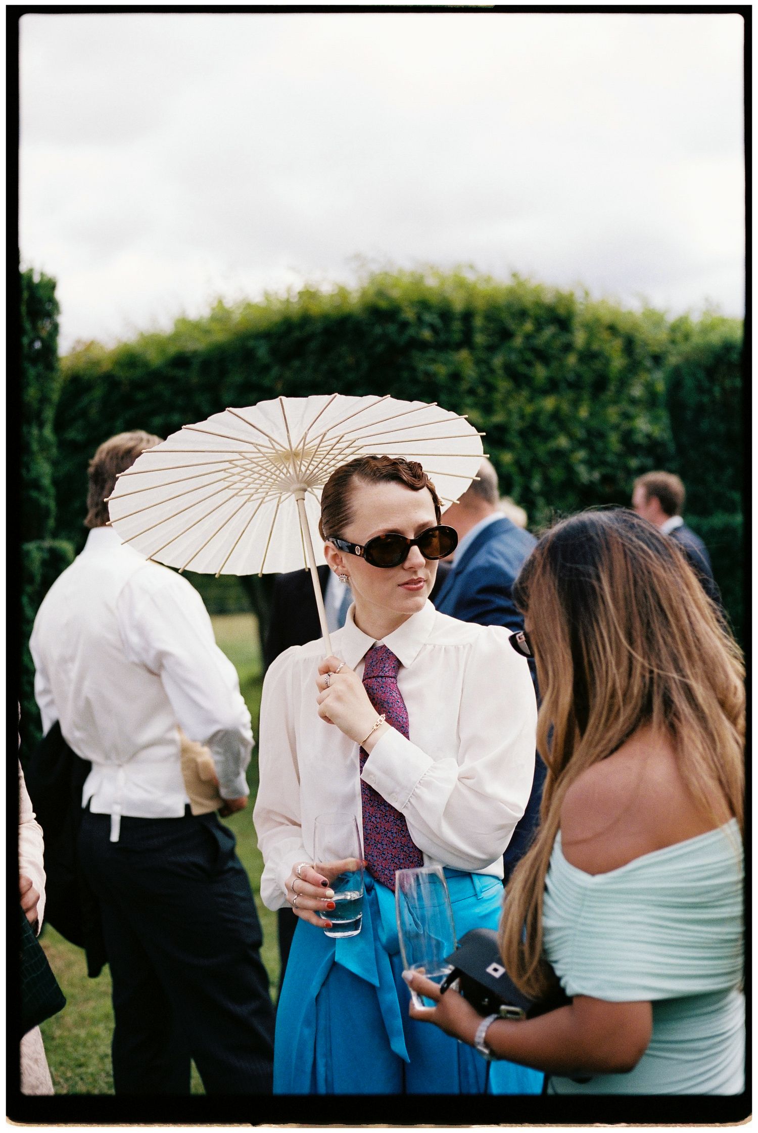 Woman in sunglasses holding a parasol and drink at an outdoor garden party wedding reception.