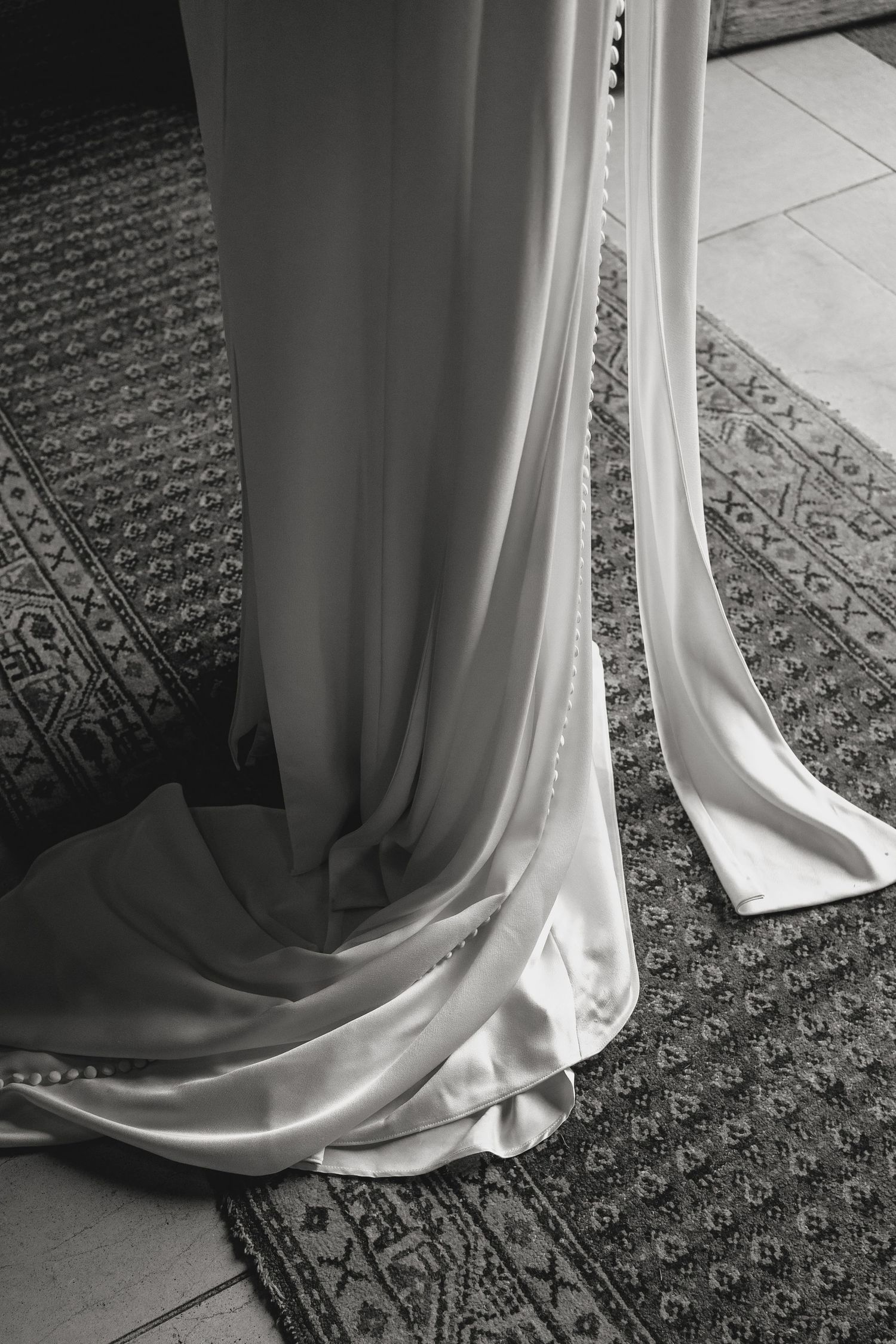 Close-up details of a white wedding dress hem trailing across an ornate patterned carpet.