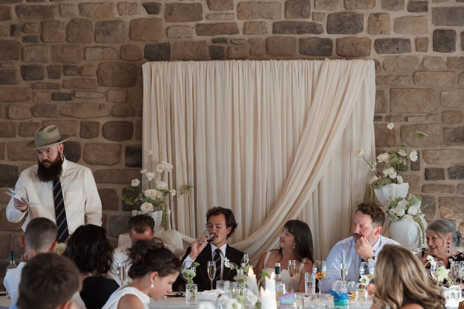 Wedding reception guests seated at decorated tables with stone wall and white curtain backdrop.