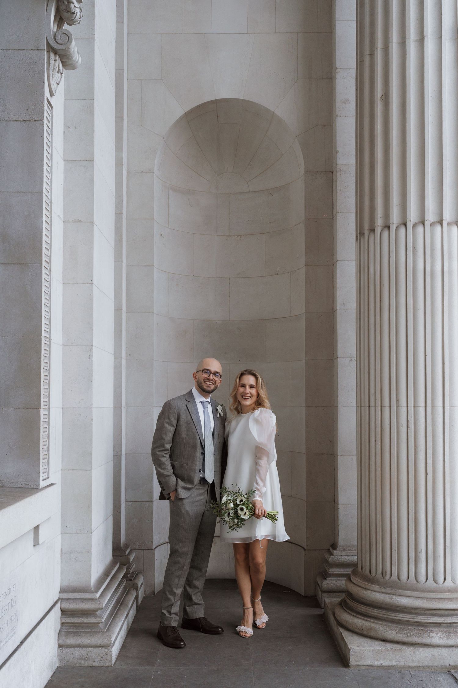 A couple posing between classical white stone columns in an elegant architectural setting.
