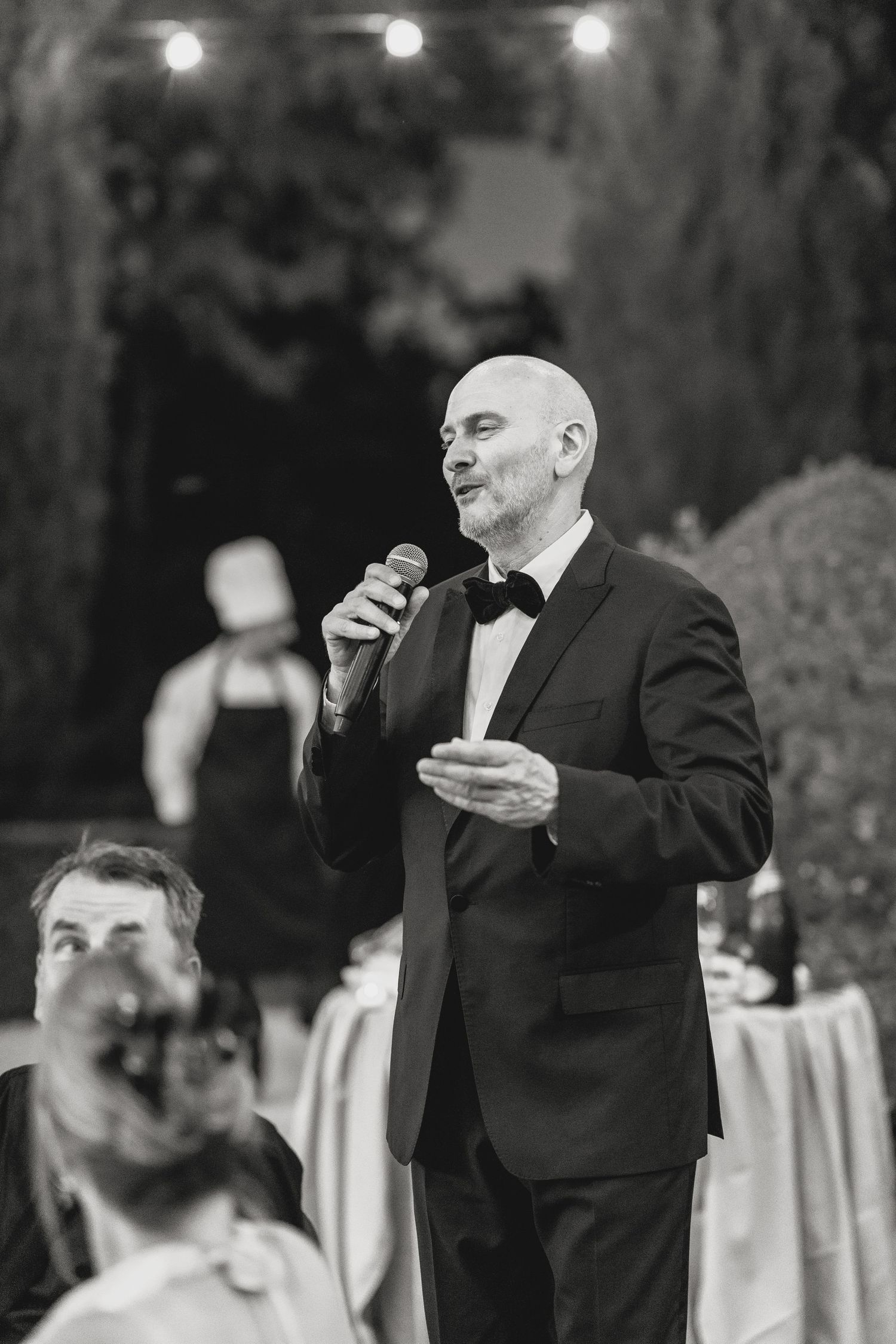 Bald man in tuxedo giving speech with microphone at formal outdoor evening event in black and white.