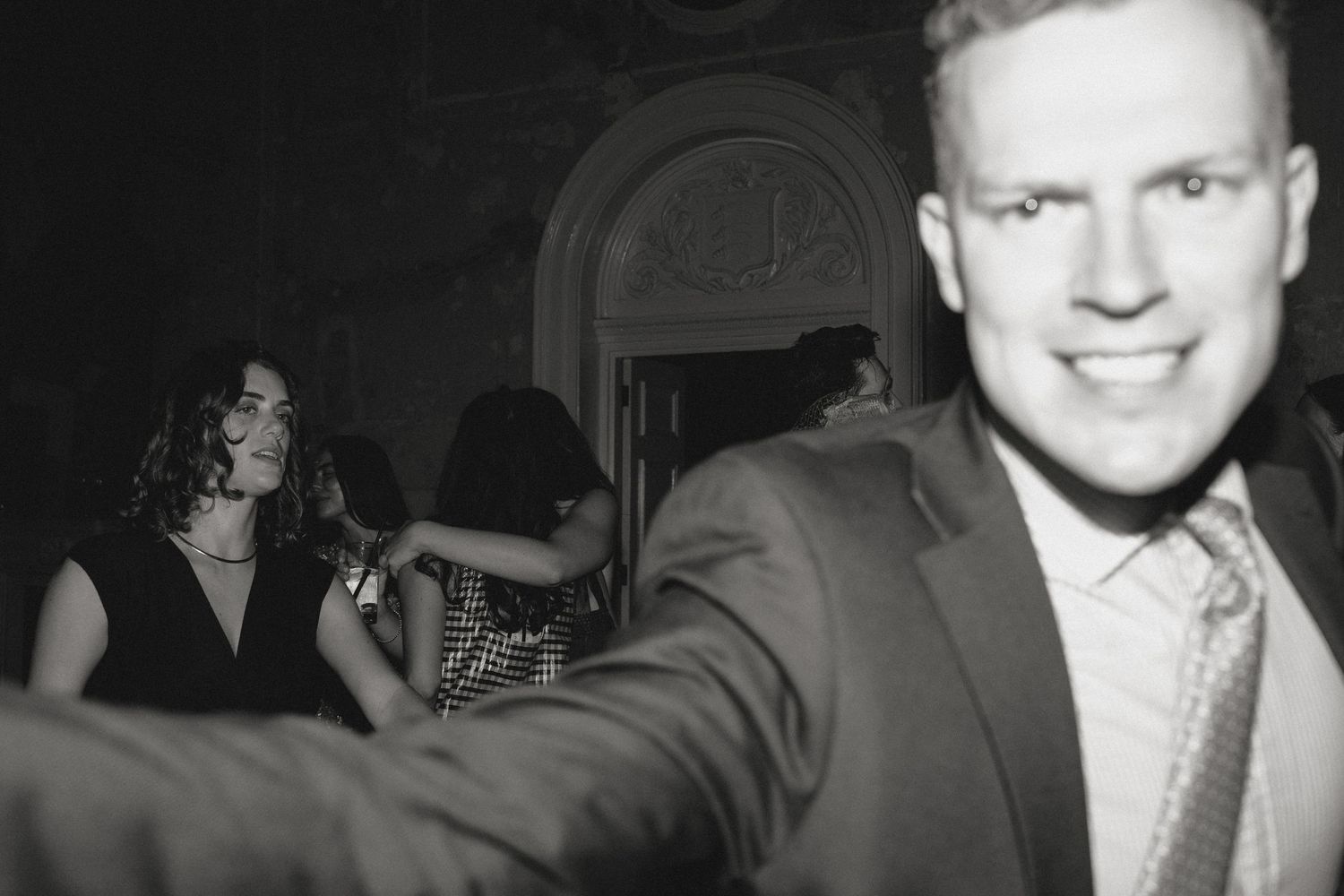 Smiling man in a suit and striped tie reaches toward the camera while guests dance in the dimly lit background of a black and white party.
