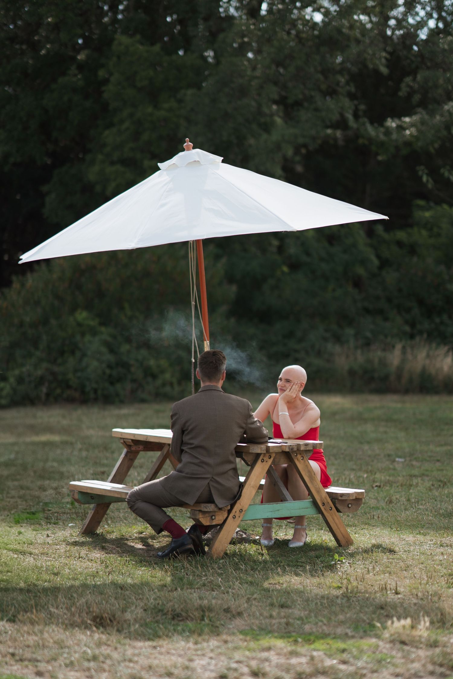 Two people sit at picnic table under white umbrella in grassy field.