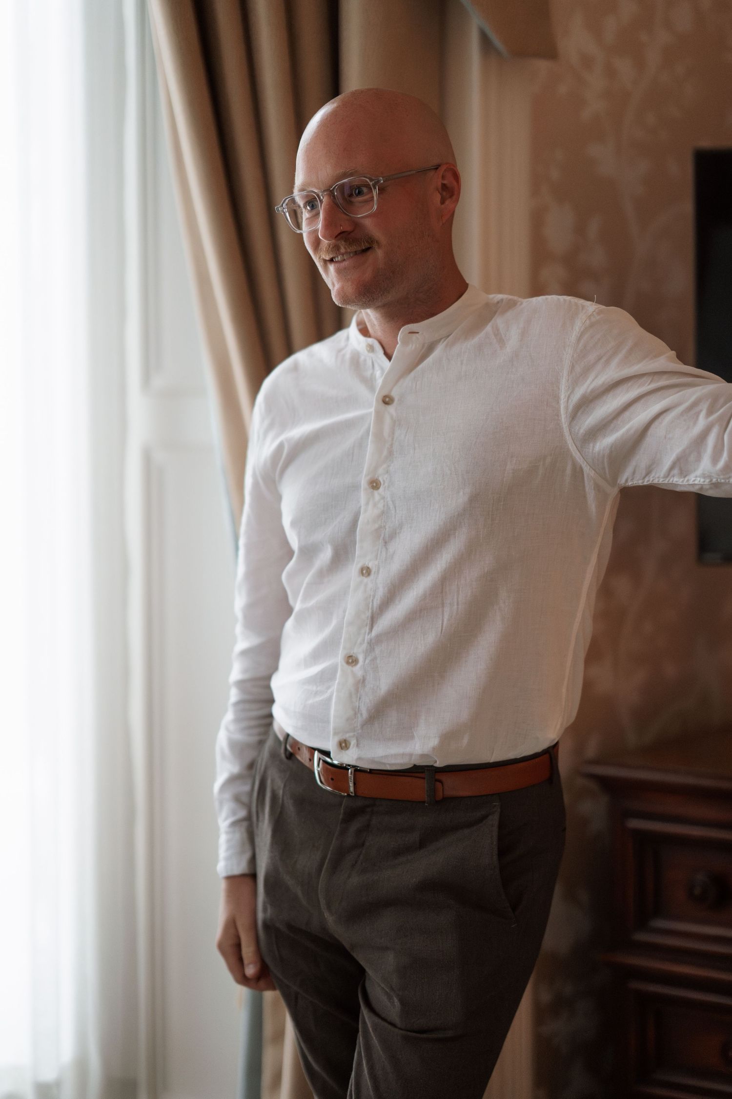 Portrait of person in white button-down shirt and gray pants standing by window.
