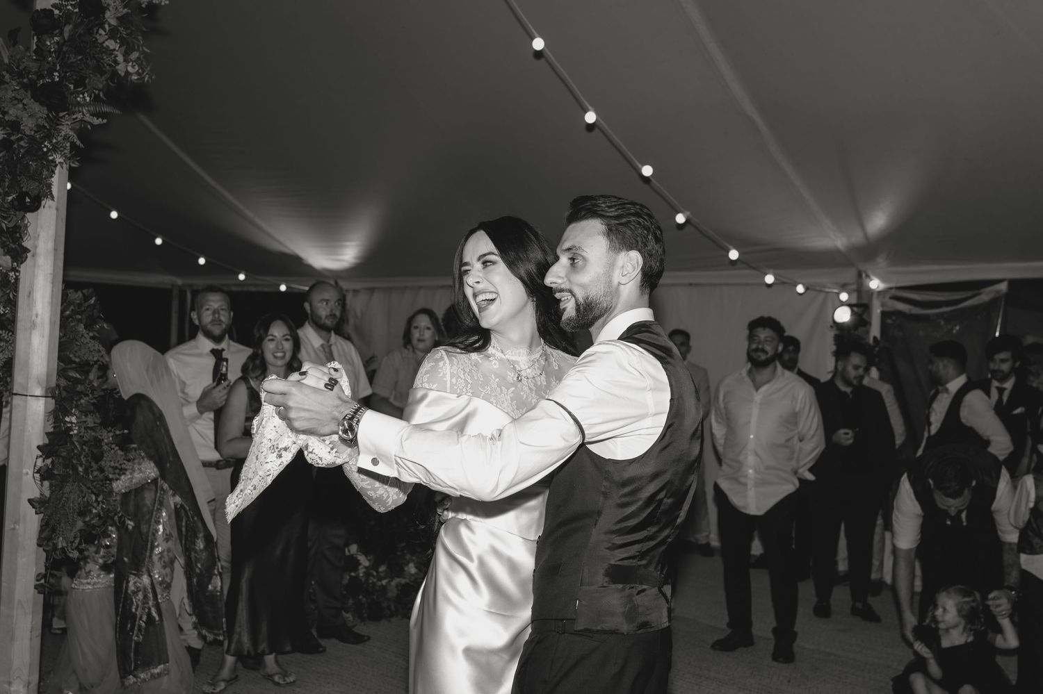 Wedding guests dancing and celebrating under string lights in a reception tent.