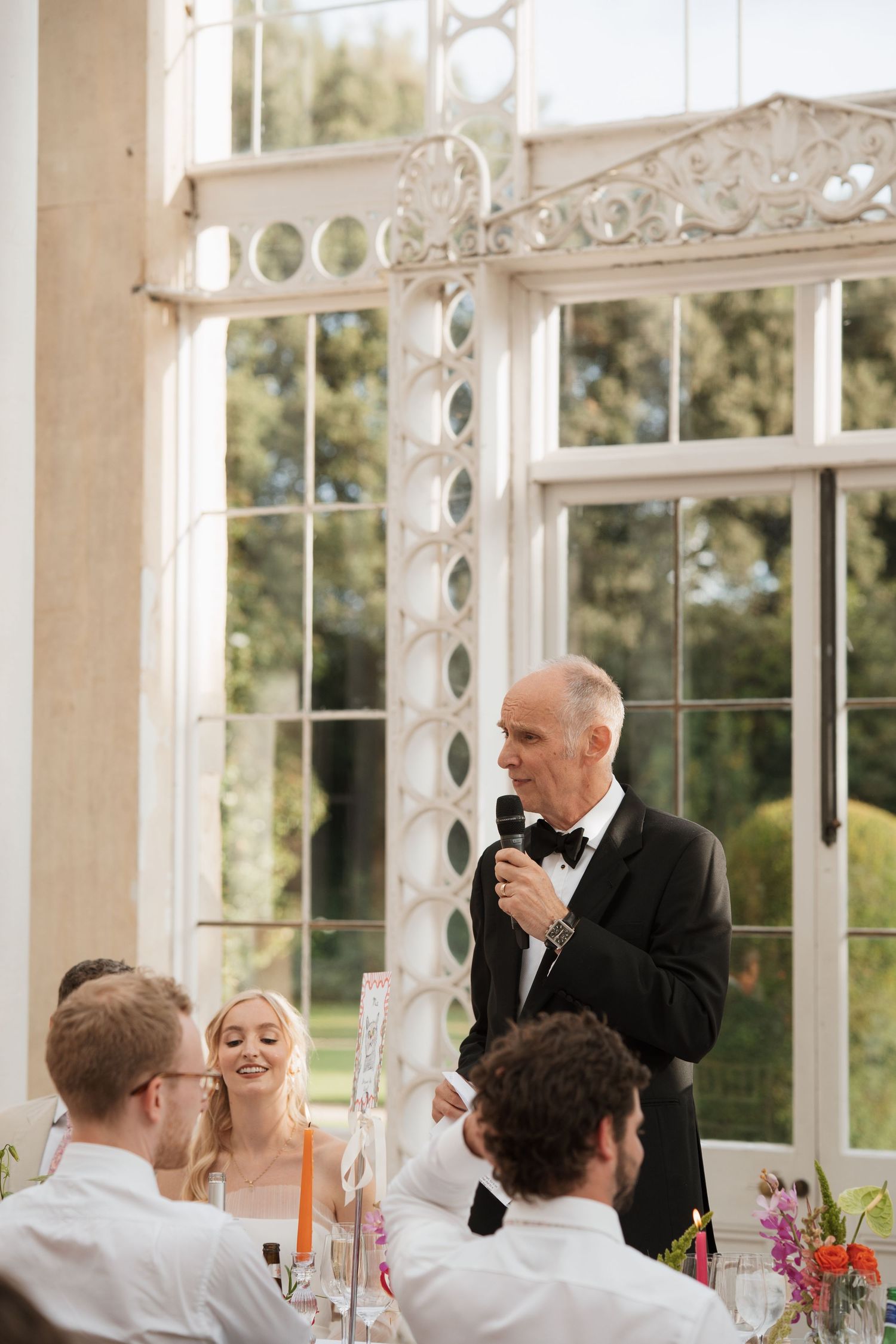 Wedding reception in a sunlit conservatory with ornate windows and guests enjoying speeches.