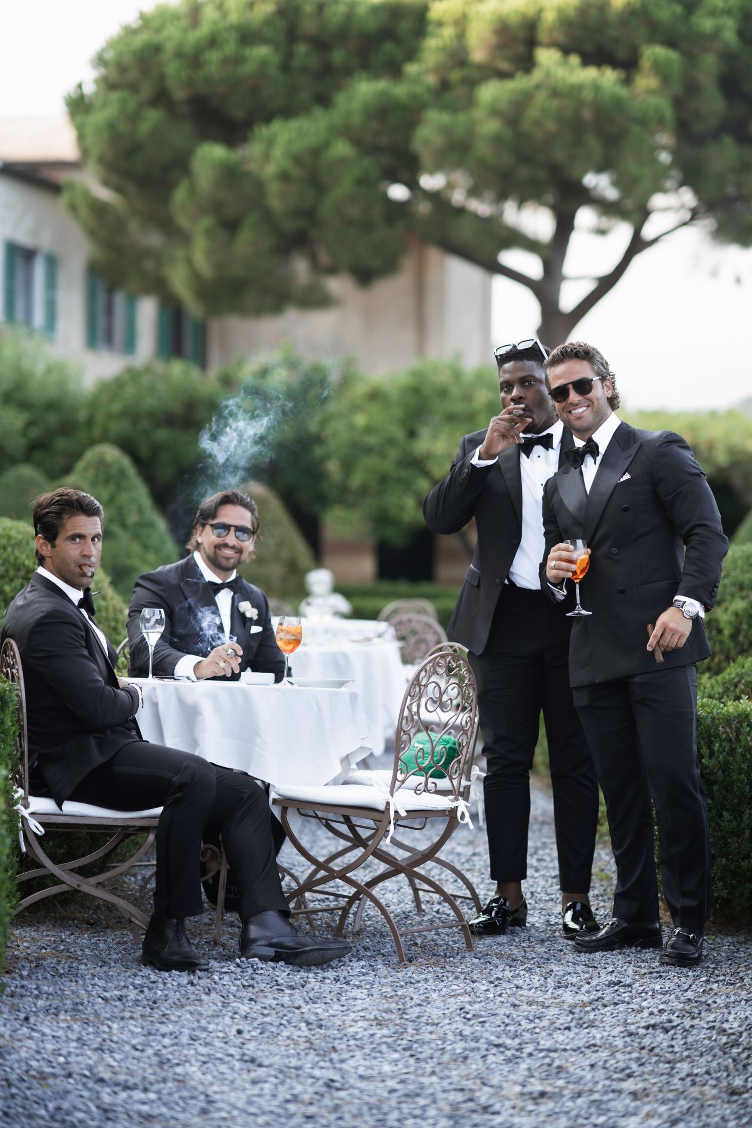 Groomsmen in black tuxedos relaxing outdoors with drinks during wedding celebration.