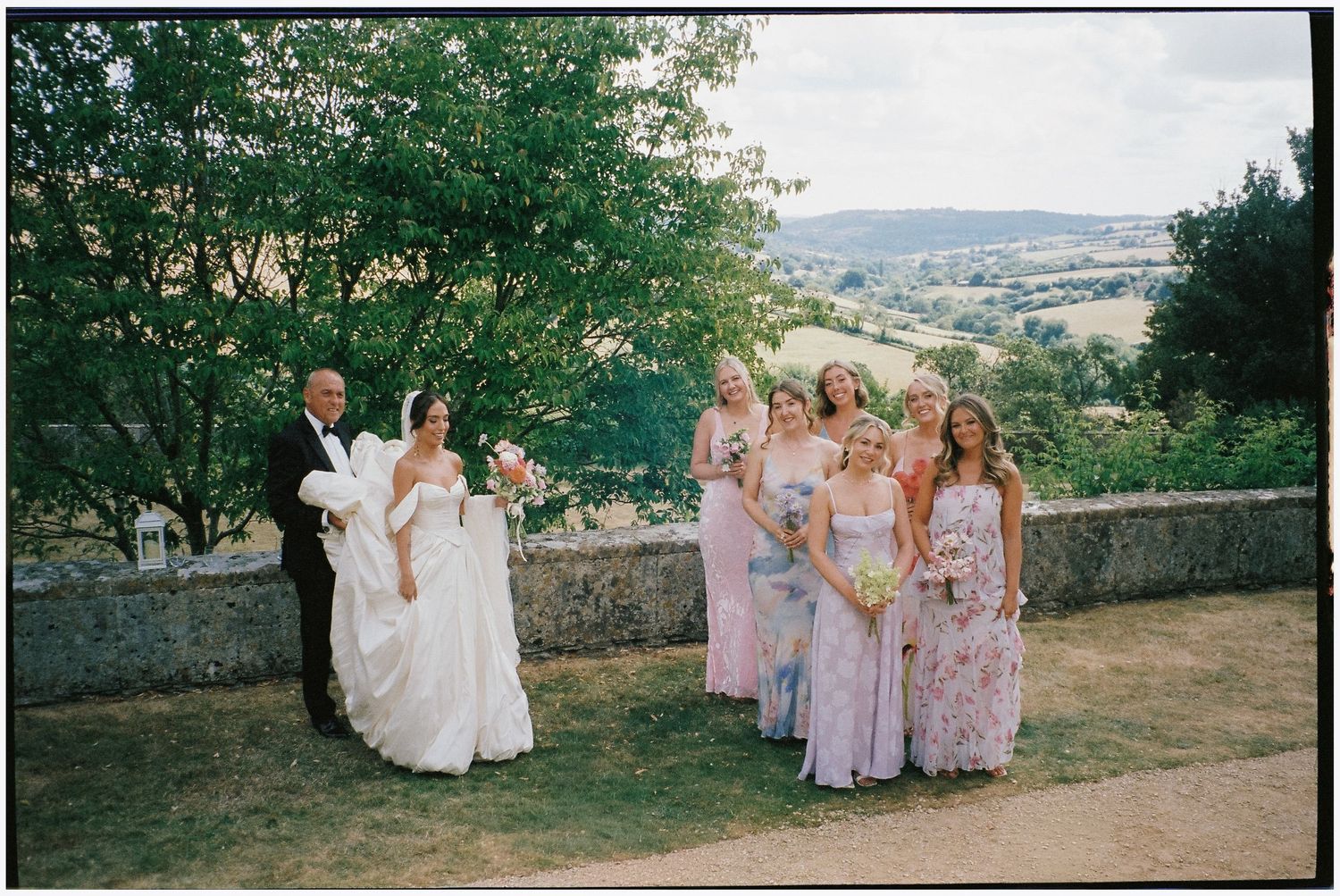 Bride in a white gown and groom in a dark suit pose beside bridesmaids in lavender dresses on a stone terrace overlooking green countryside.