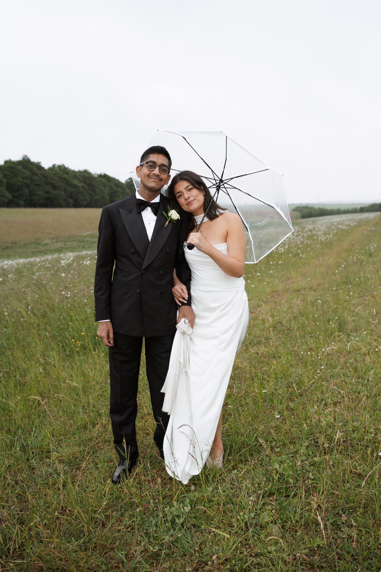 Wedding couple poses together in a meadow with a clear umbrella against overcast sky.