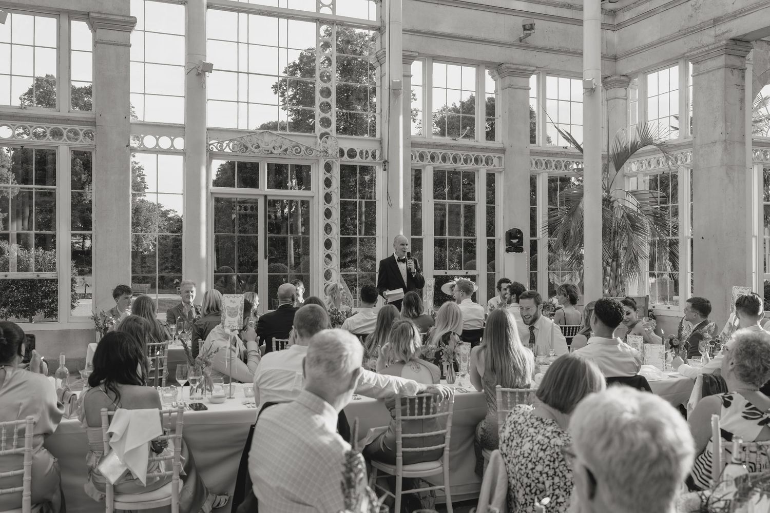 Black and white photo of wedding guests dining in a bright conservatory with large windows.