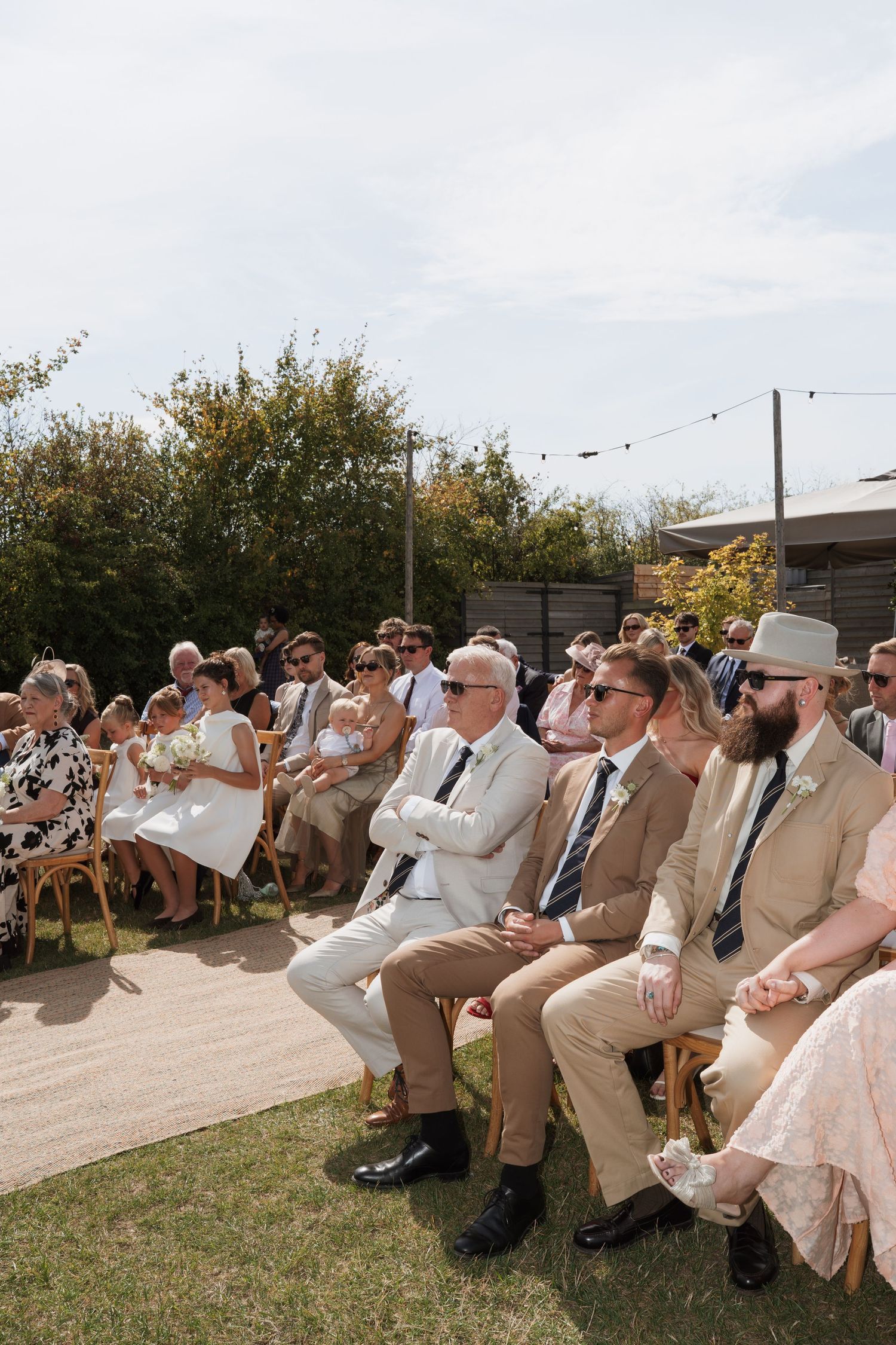 Wedding guests in coordinated beige attire seated in rows during outdoor ceremony.