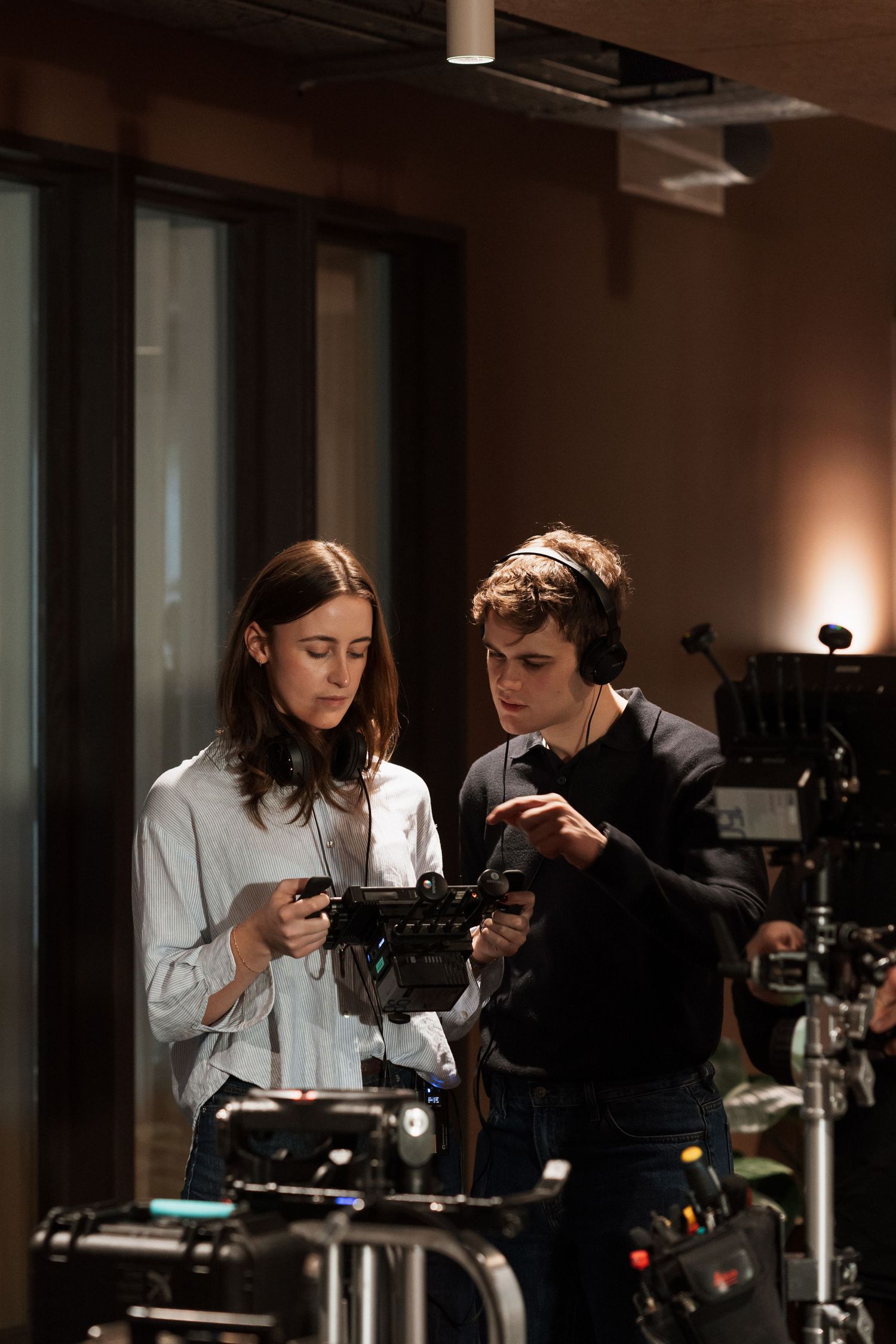 Two filmmakers review footage on a professional camera on a dimly lit film set with equipment visible.