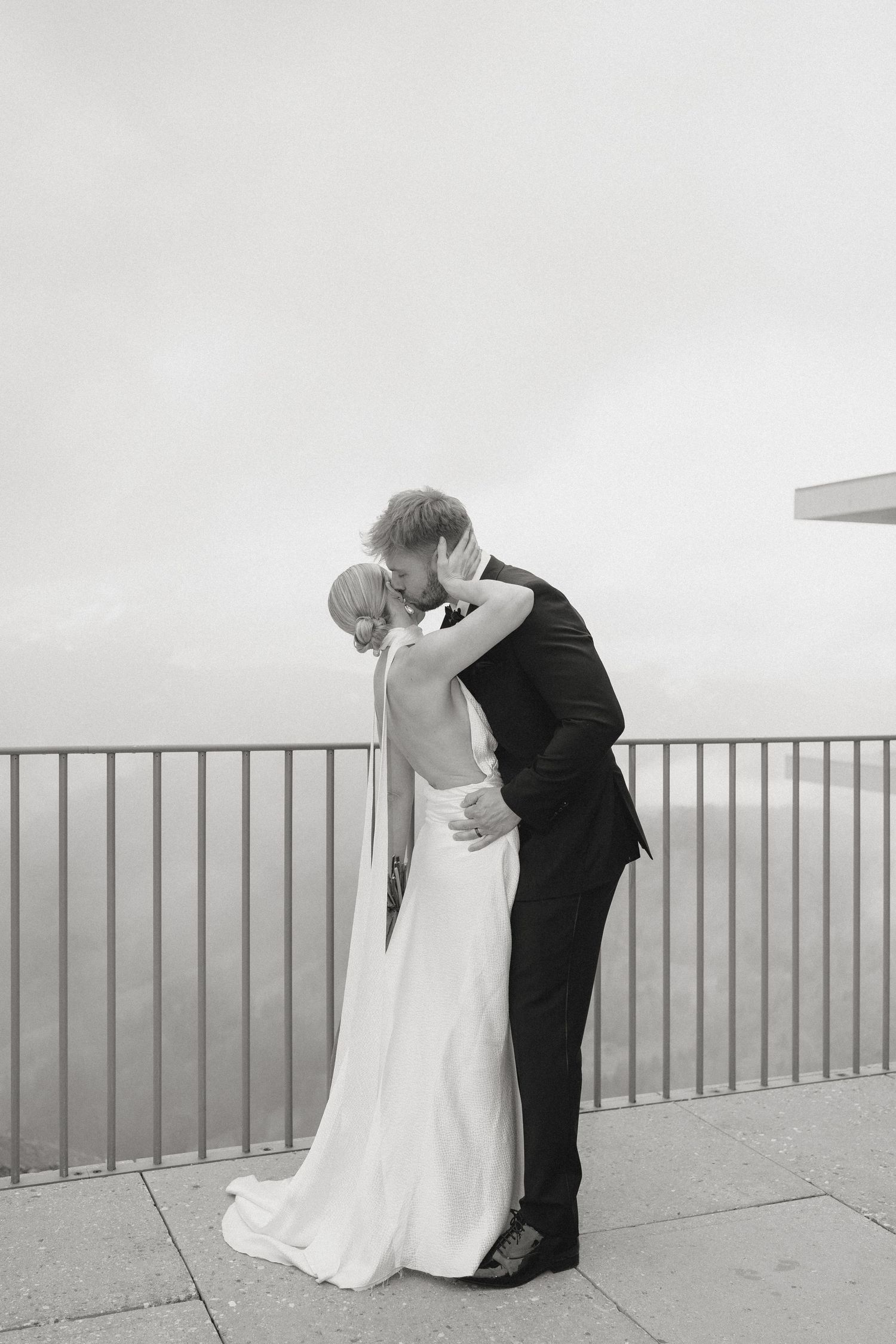 Black and white photo of a couple in wedding attire on a balcony overlooking the ocean.