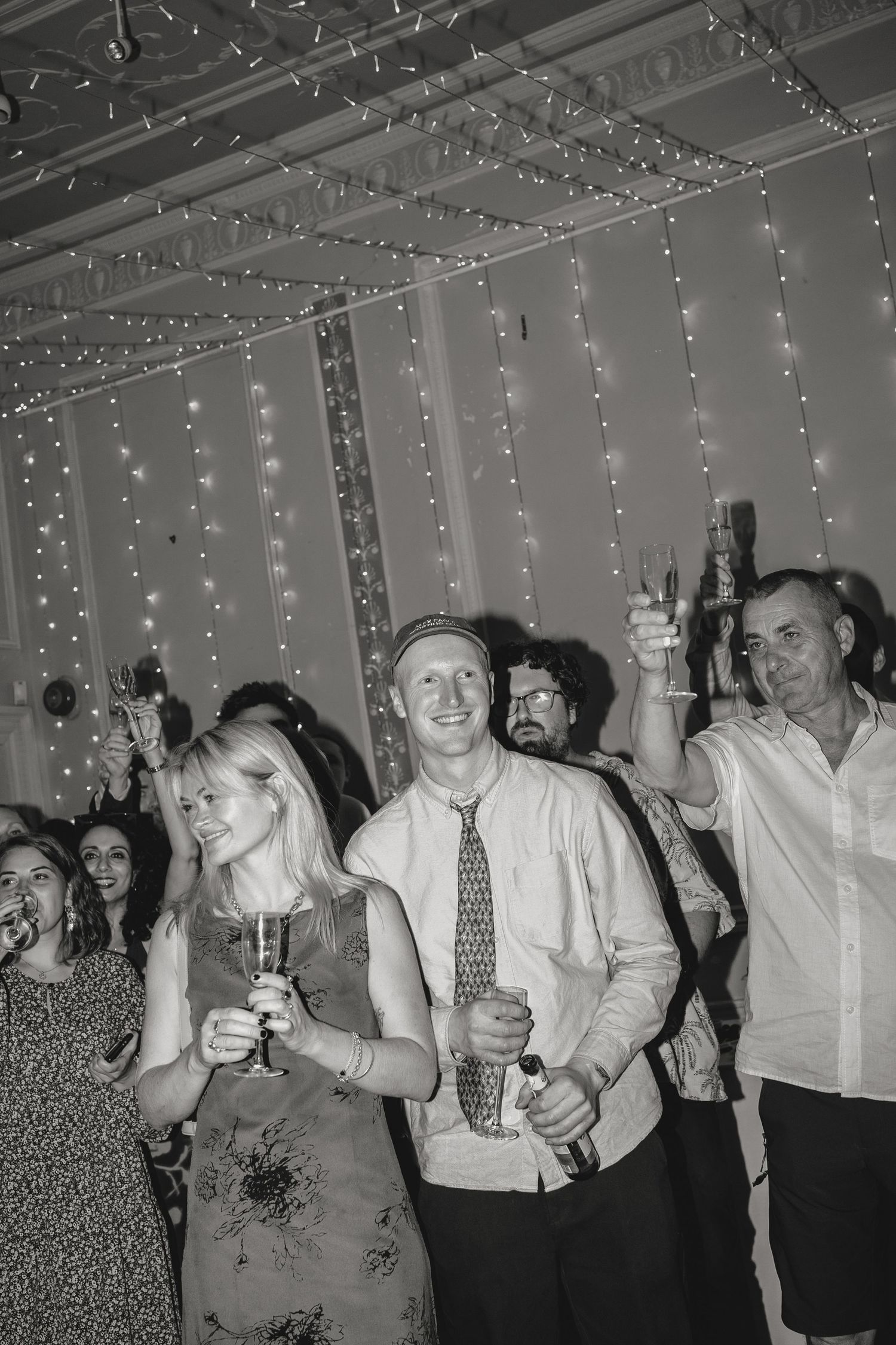 Wedding guests raising their hands and drinks in celebration under string lights.